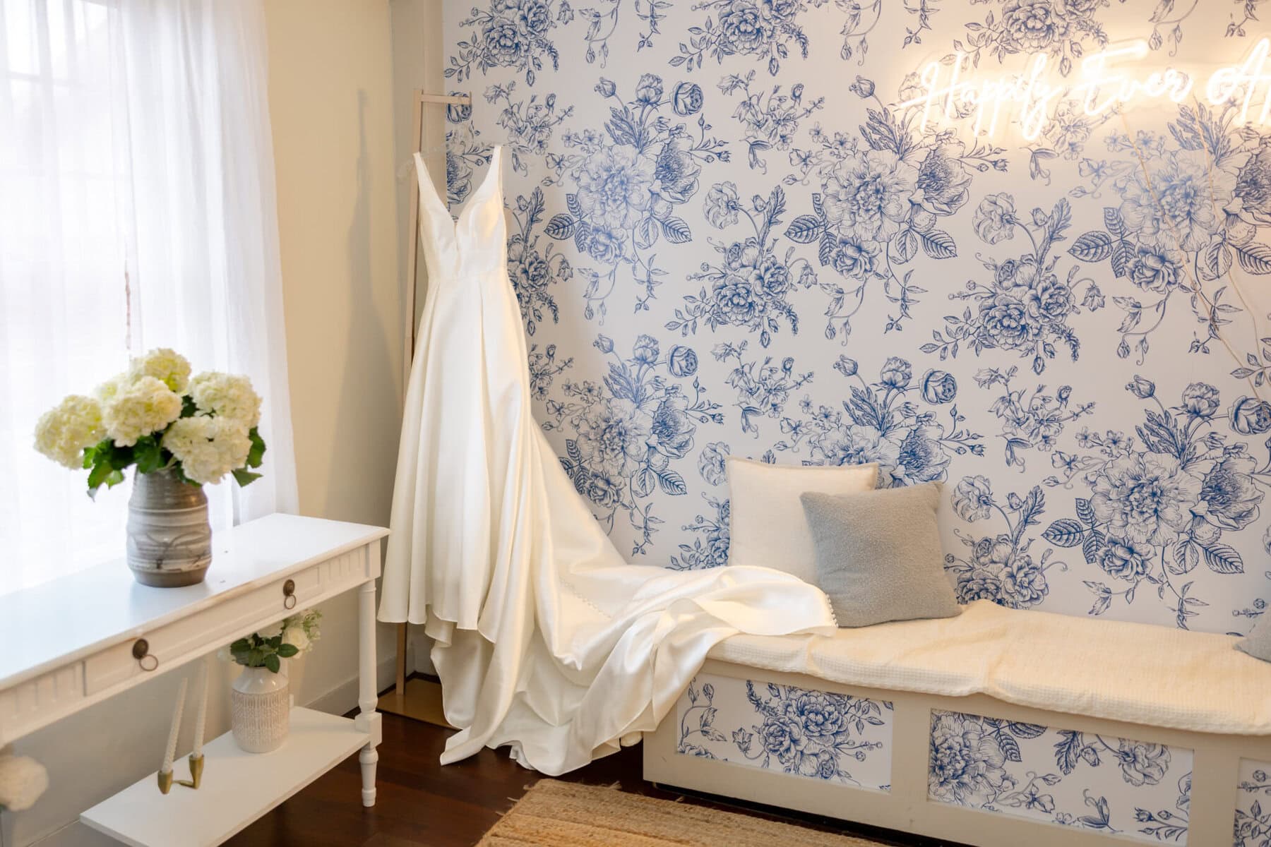Wedding Reception at the Barker House 3 A long white wedding dress hangs in Barker House, beside a cushioned bench and white flowers—perfect for a memorable wedding reception.