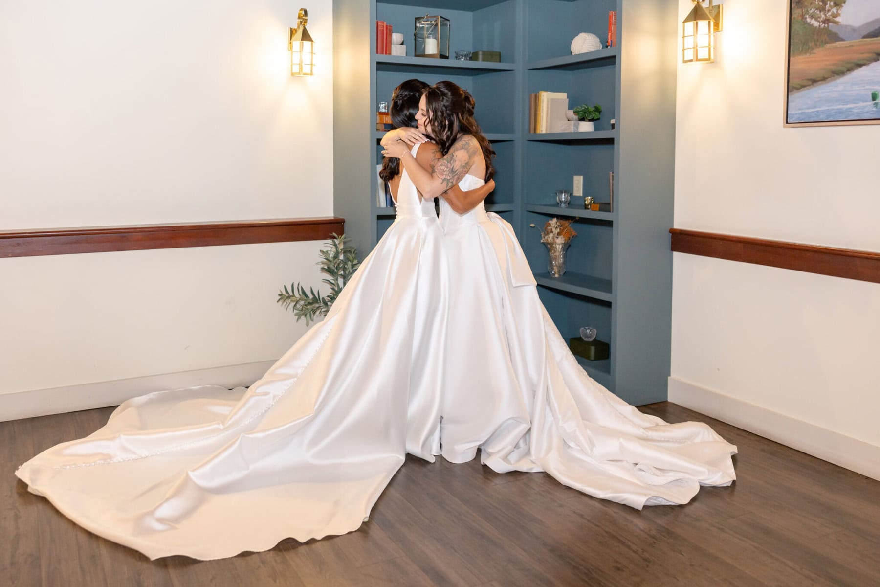 Wedding Reception at the Barker House 11 Two brides in white gowns embrace by a blue bookshelf at their Barker House wedding reception, lit by wall sconces and natural light.