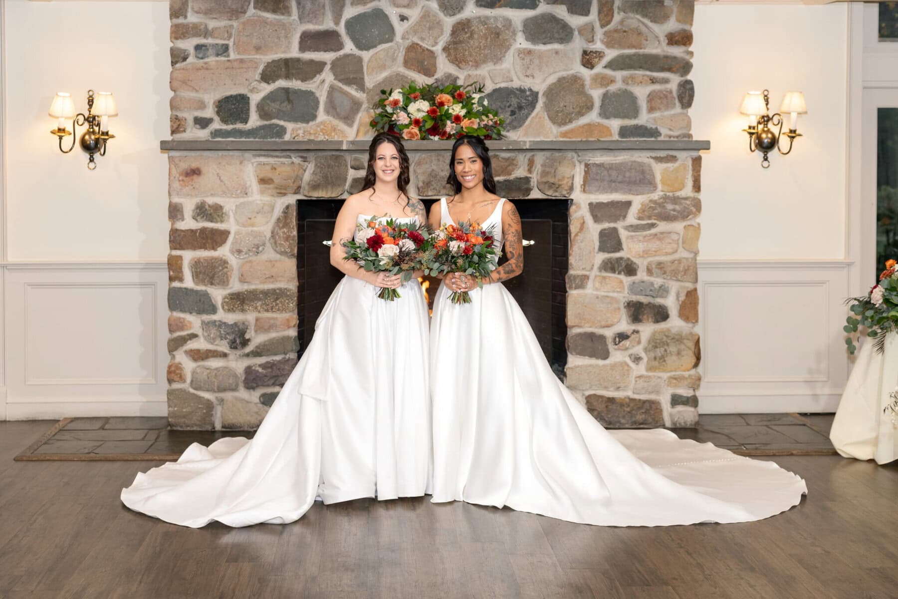 Wedding Reception at the Barker House 14 Two brides in white gowns stand holding bouquets at Barker House, ready to celebrate their wedding reception by a flower-adorned stone fireplace.