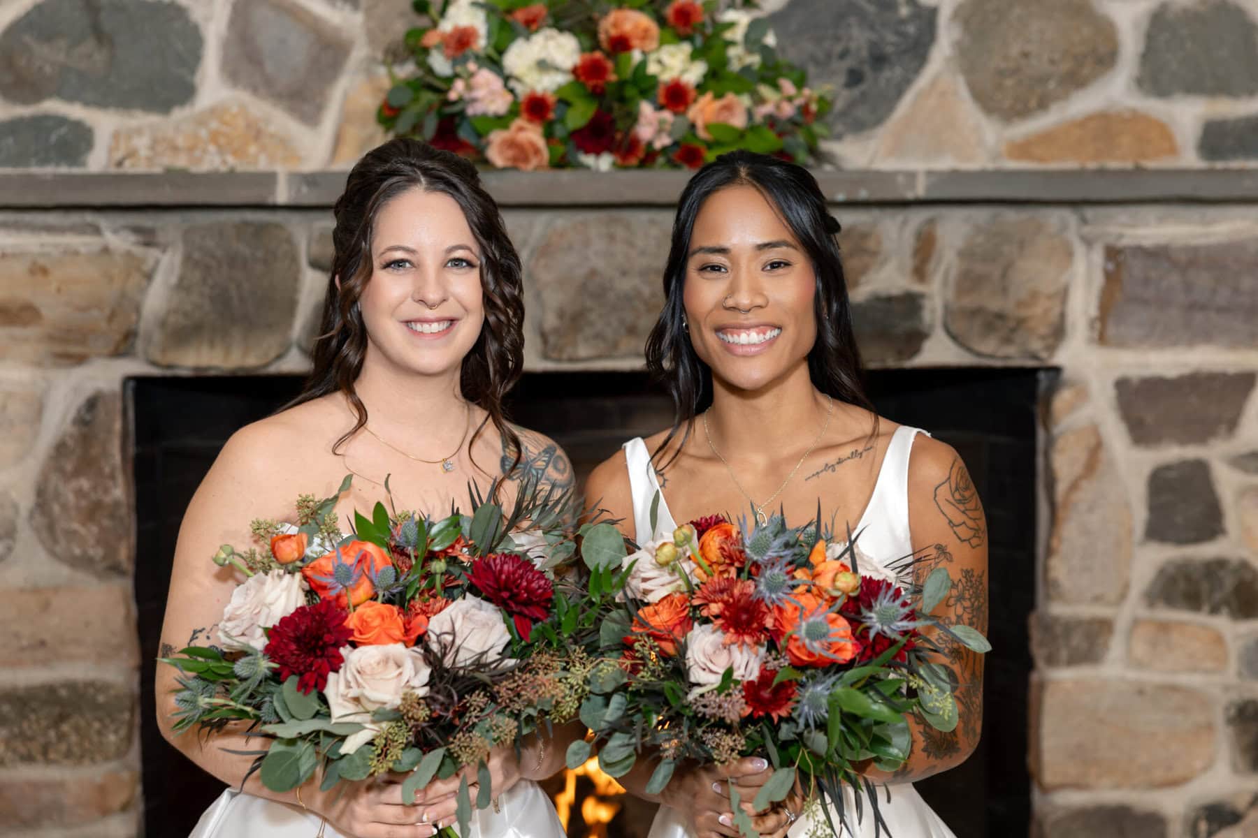 Wedding Reception at the Barker House 16 Two women in white dresses hold colorful floral bouquets at the Barker House, standing before a stone fireplace at a wedding reception.