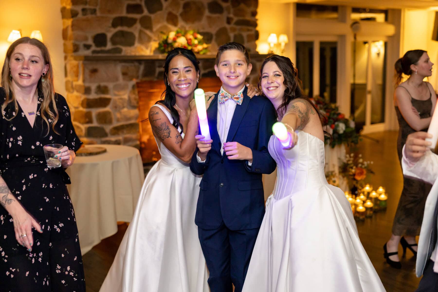 Wedding Reception at the Barker House 31 Two women in wedding dresses and a boy in a suit smile at a Barker House wedding reception, holding glow sticks with others nearby.