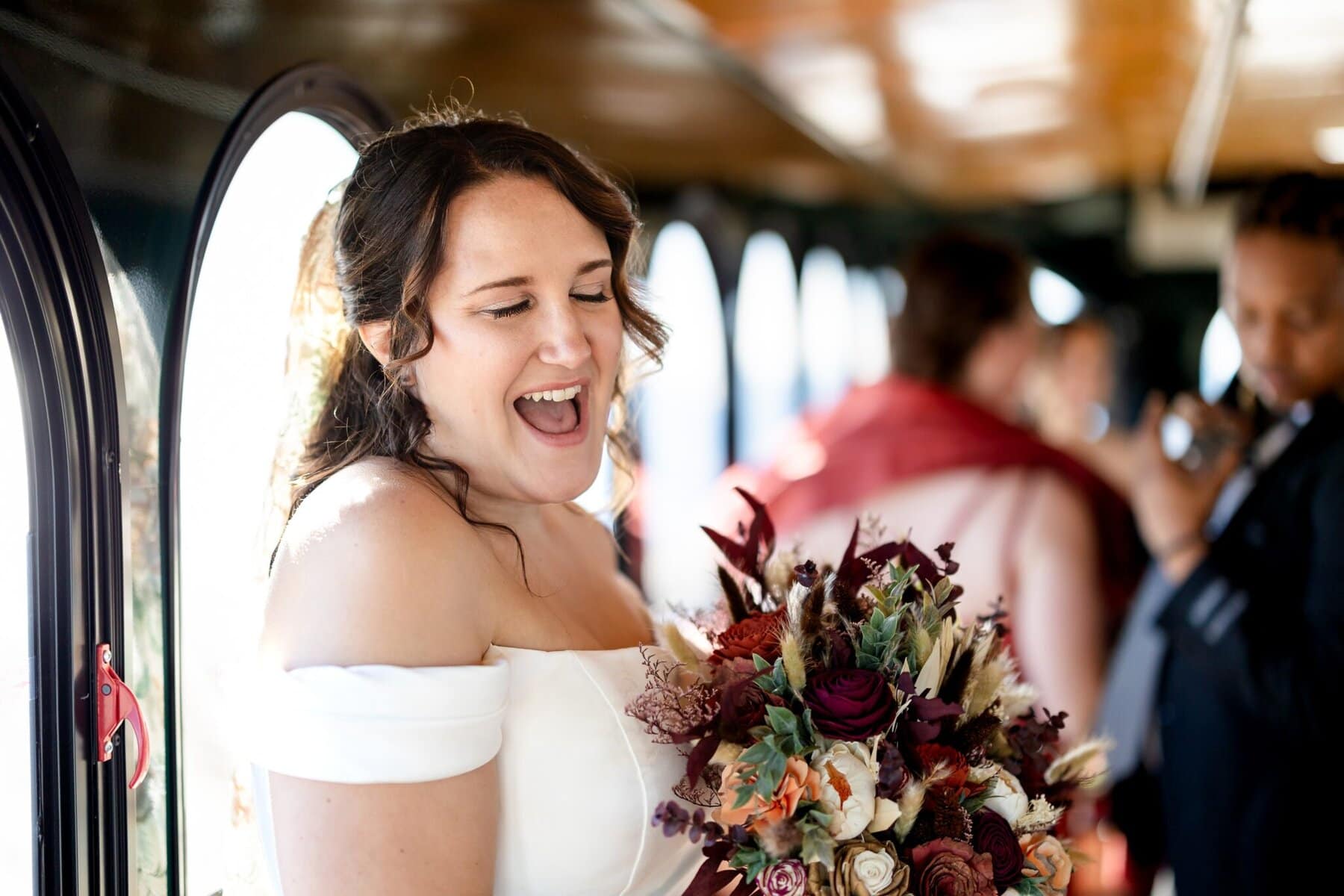 Wedding at the Newport Naval Station Officers' Club 8 A bride in an off-shoulder dress beams, holding her bouquet inside a sunlit vehicle at Newport Naval Station with friends in the background.