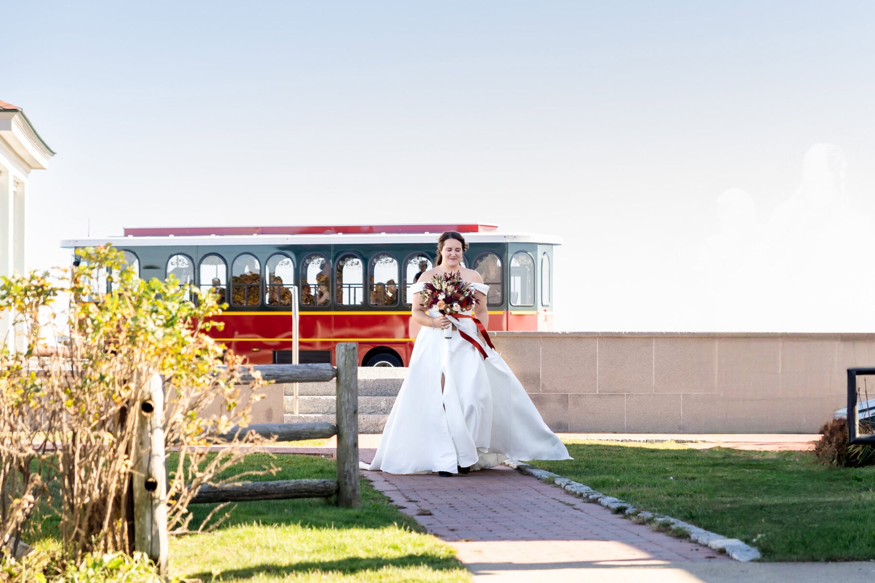 Wedding at the Newport Naval Station Officers' Club 13 A bride in a white dress stands on a walkway outside the Officers' Club, holding a bouquet, with a red trolley visible in the background.