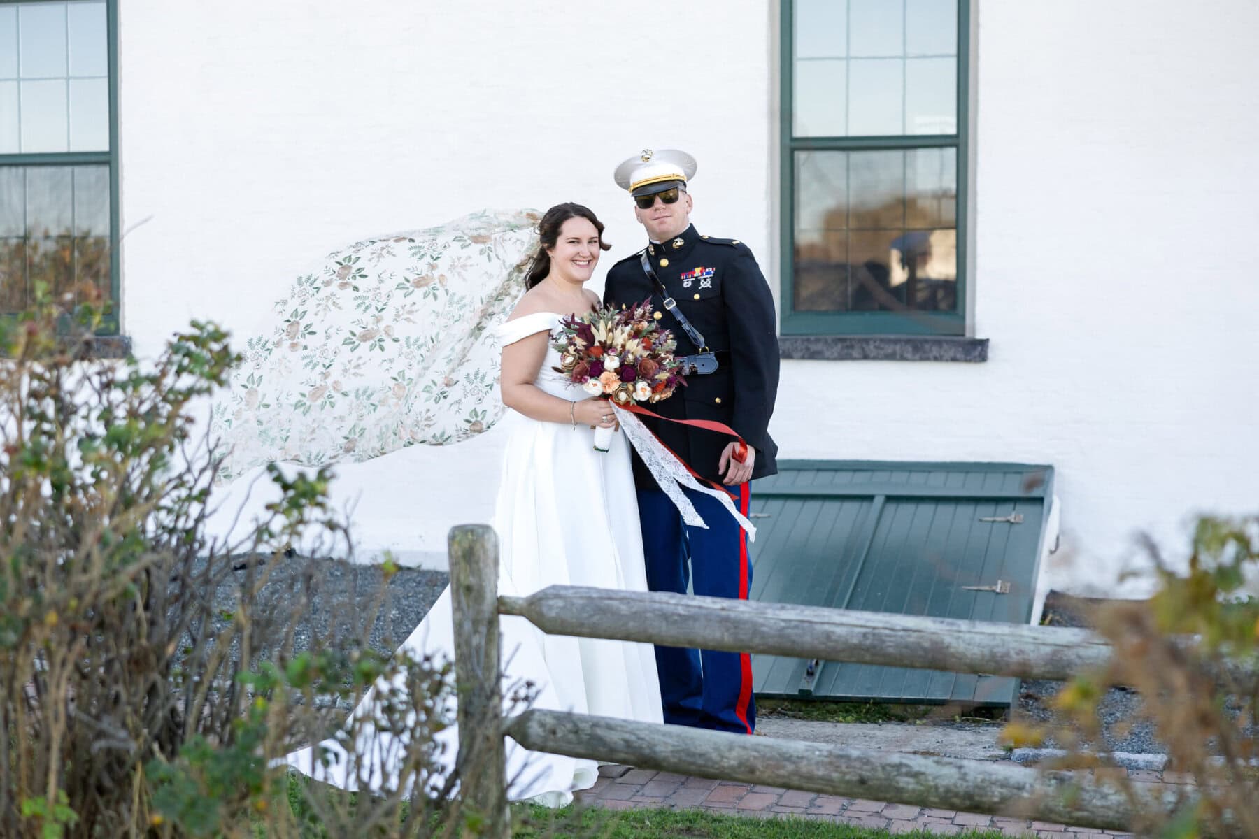 Wedding at the Newport Naval Station Officers' Club 21 A bride in white and a person in uniform pose outside by a fence, celebrating their Newport Naval Station wedding.