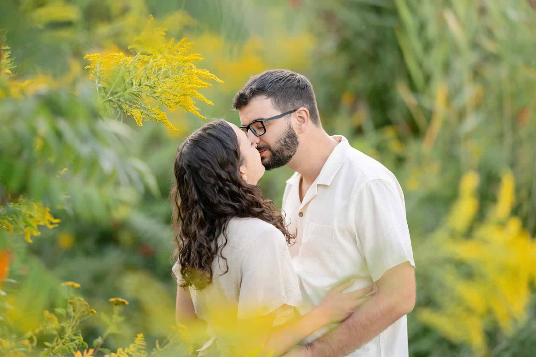 Webb Park in Weymouth, MA Engagement Session 9 A couple stands close, sharing a gentle kiss in lush greenery and yellow flowers at Webb Park, Weymouth MA, both wearing light-colored shirts during their engagement session.