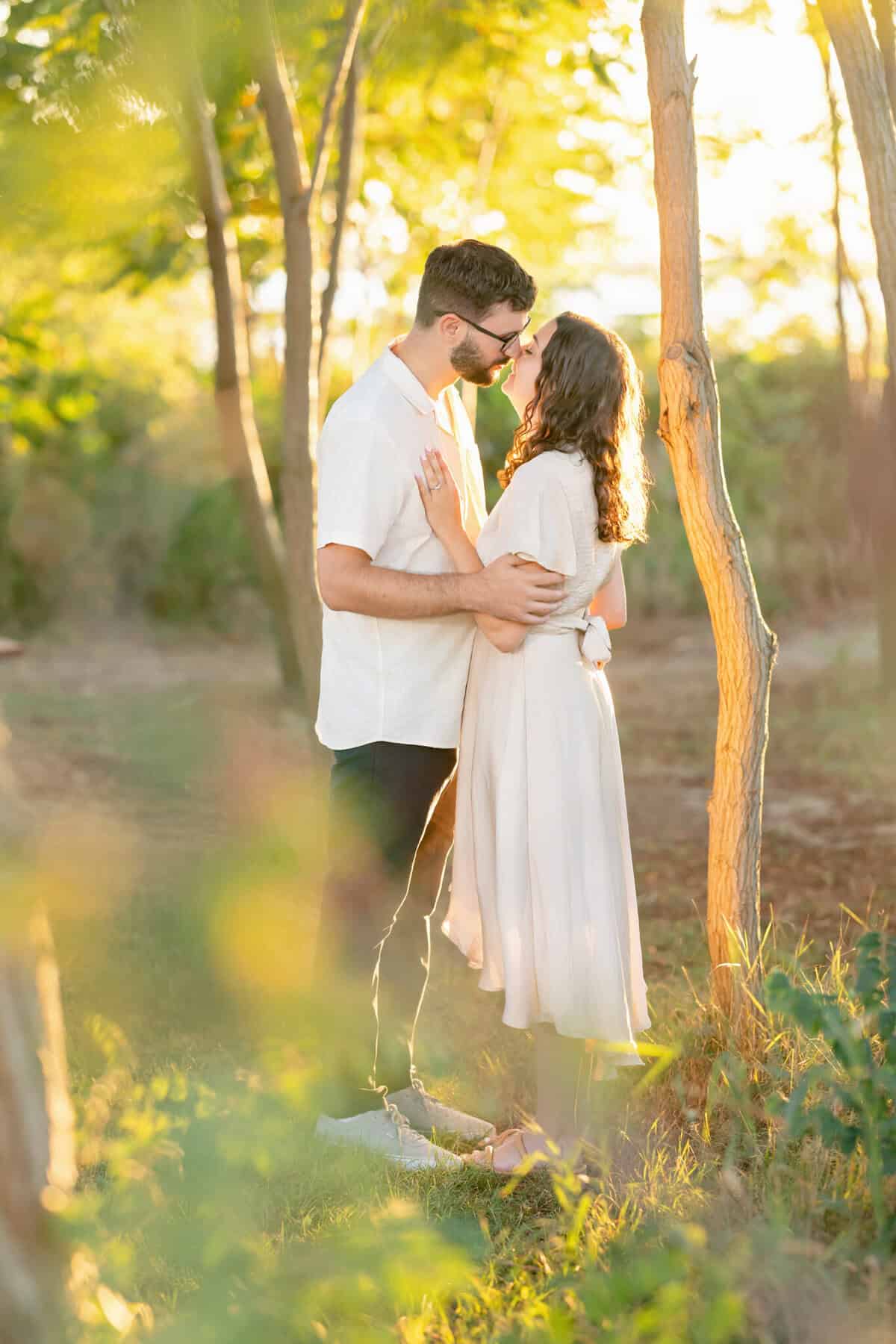 Webb Park in Weymouth, MA Engagement Session 16 A couple stands closely in sunlit Webb Park, Weymouth MA, sharing gentle smiles before a kiss—perfect for an engagement session. She wears a light dress; he’s in a white shirt and dark pants.