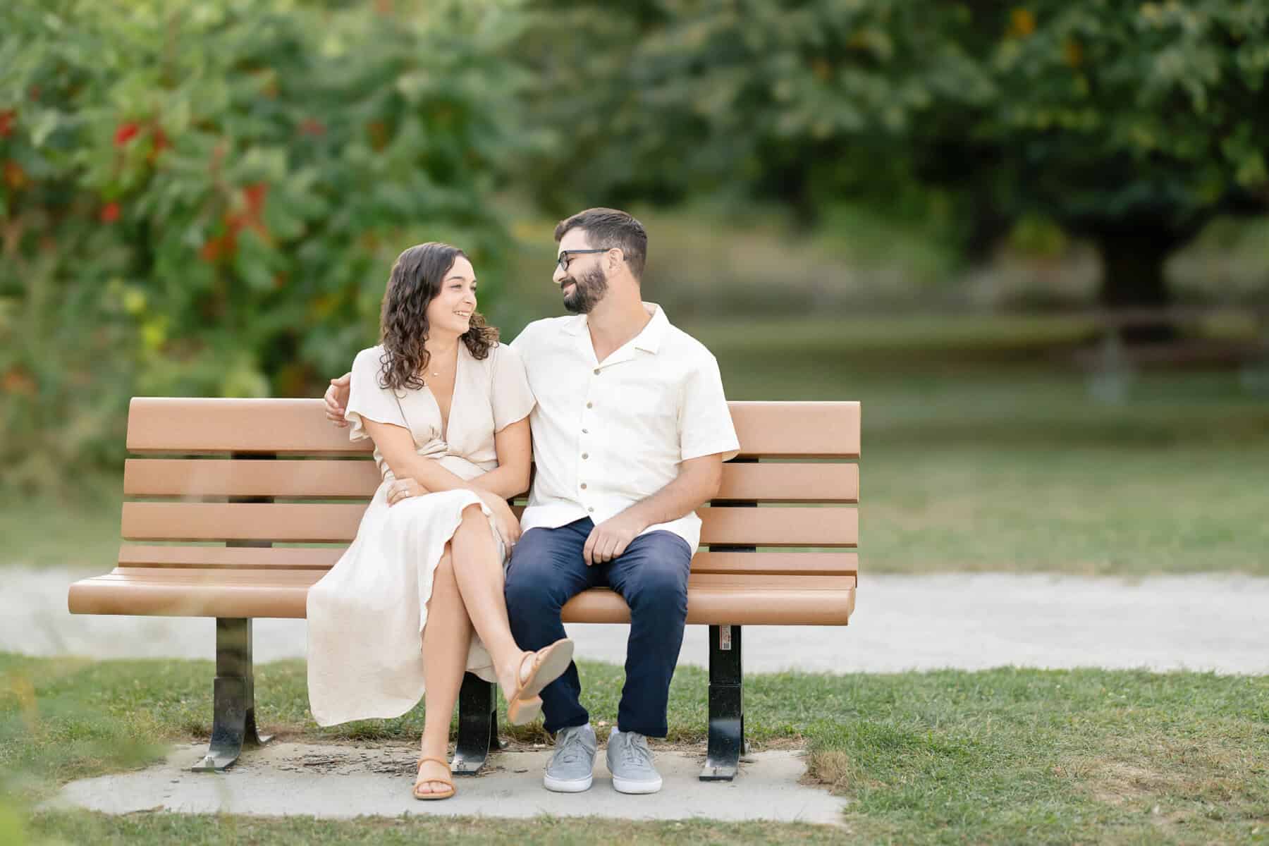 Webb Park in Weymouth, MA Engagement Session 6 A smiling couple sits closely on a park bench in Webb Park, Weymouth MA, both wearing light clothes, gazing at each other amid greenery—perfect for an engagement session.
