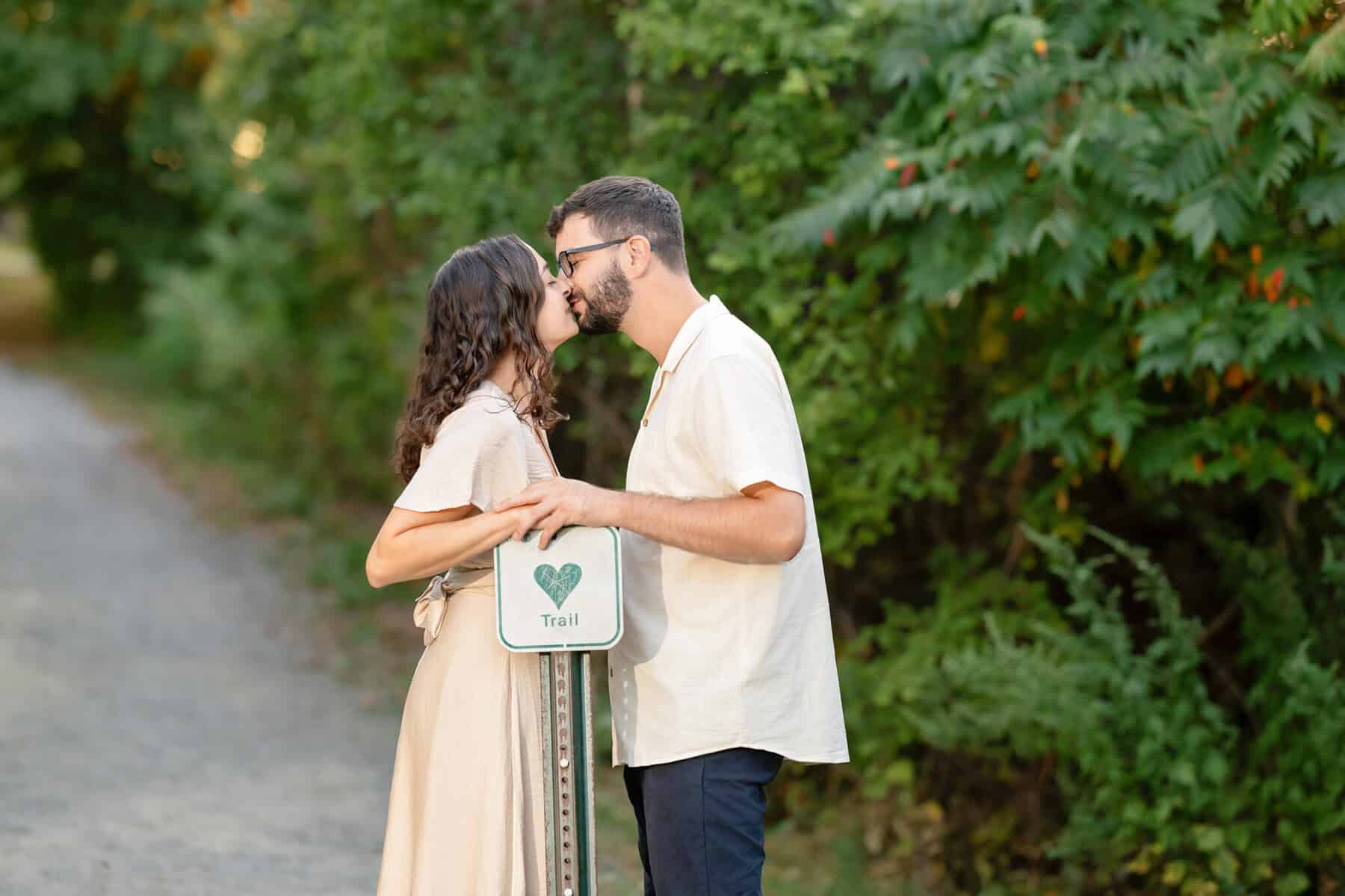 Webb Park in Weymouth, MA Engagement Session 18 A couple stands on a wooded trail in Webb Park, touching foreheads affectionately and holding a heart “Trail” sign, surrounded by greenery during their engagement session.