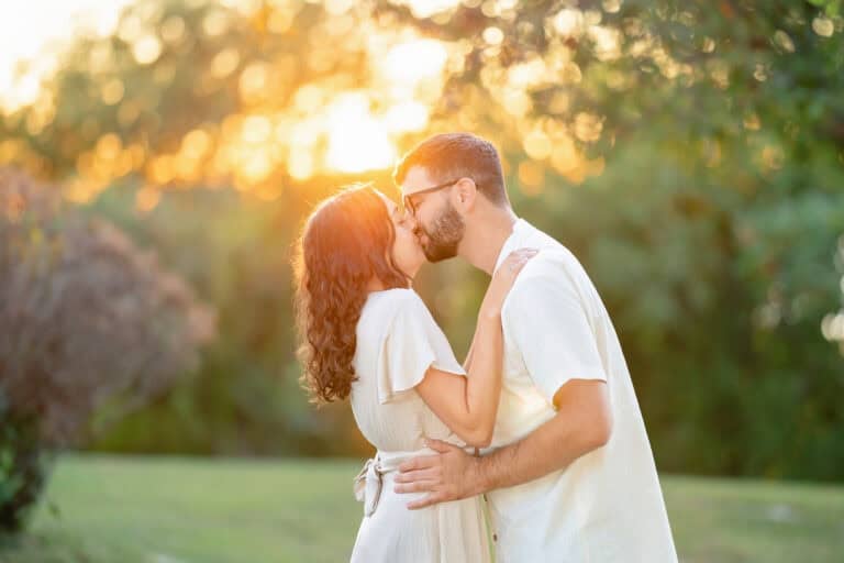 A couple in light clothing kisses at sunset in Webb Park, Weymouth MA, during a romantic engagement session surrounded by lush greenery and warm sunlight.
