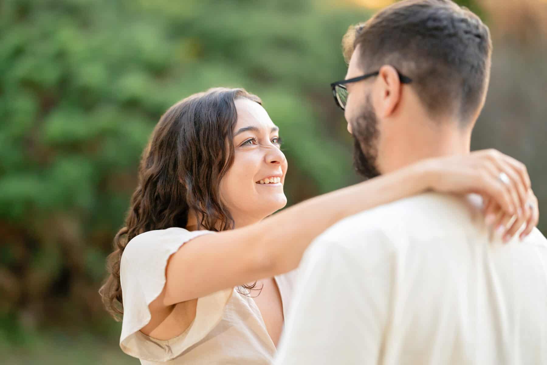 Webb Park in Weymouth, MA Engagement Session 25 A woman smiles and embraces a bearded man outdoors at Webb Park in Weymouth MA during their engagement session, both in light clothes with greenery blurred in the background.
