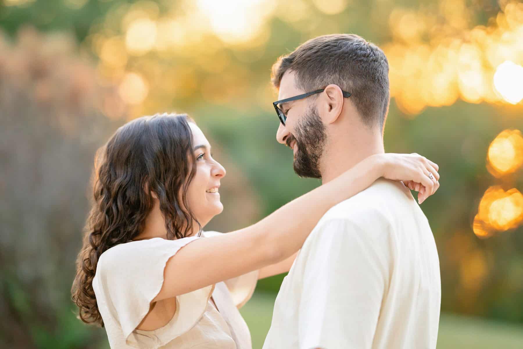 Webb Park in Weymouth, MA Engagement Session 27 A smiling woman with long curly hair embraces a bearded man in glasses during their Webb Park engagement session, surrounded by soft golden sunlight and blurred greenery in Weymouth MA.
