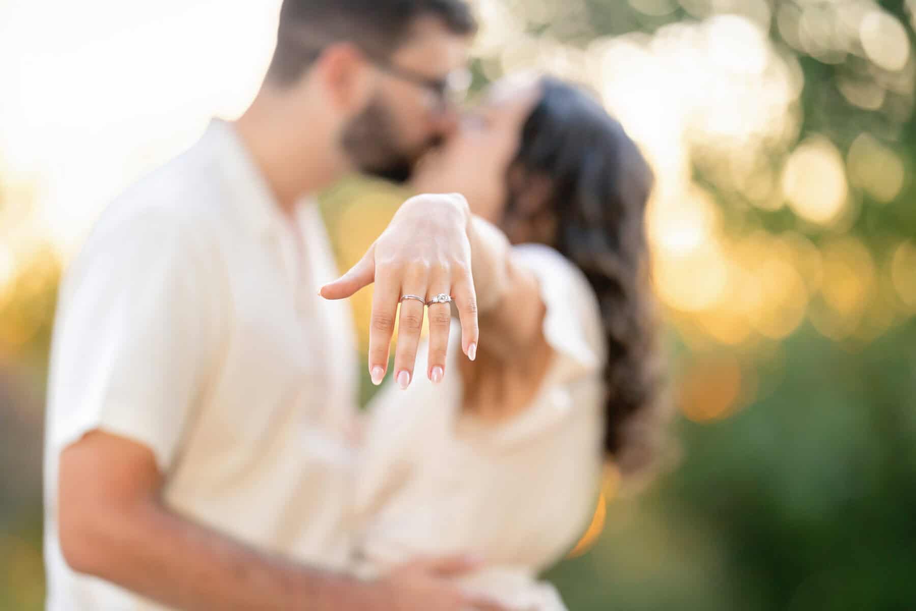 Webb Park in Weymouth, MA Engagement Session 28 A couple kisses softly in the background during their engagement session at Webb Park, as the woman extends her hand toward the camera, showcasing her ring in warm, natural light.