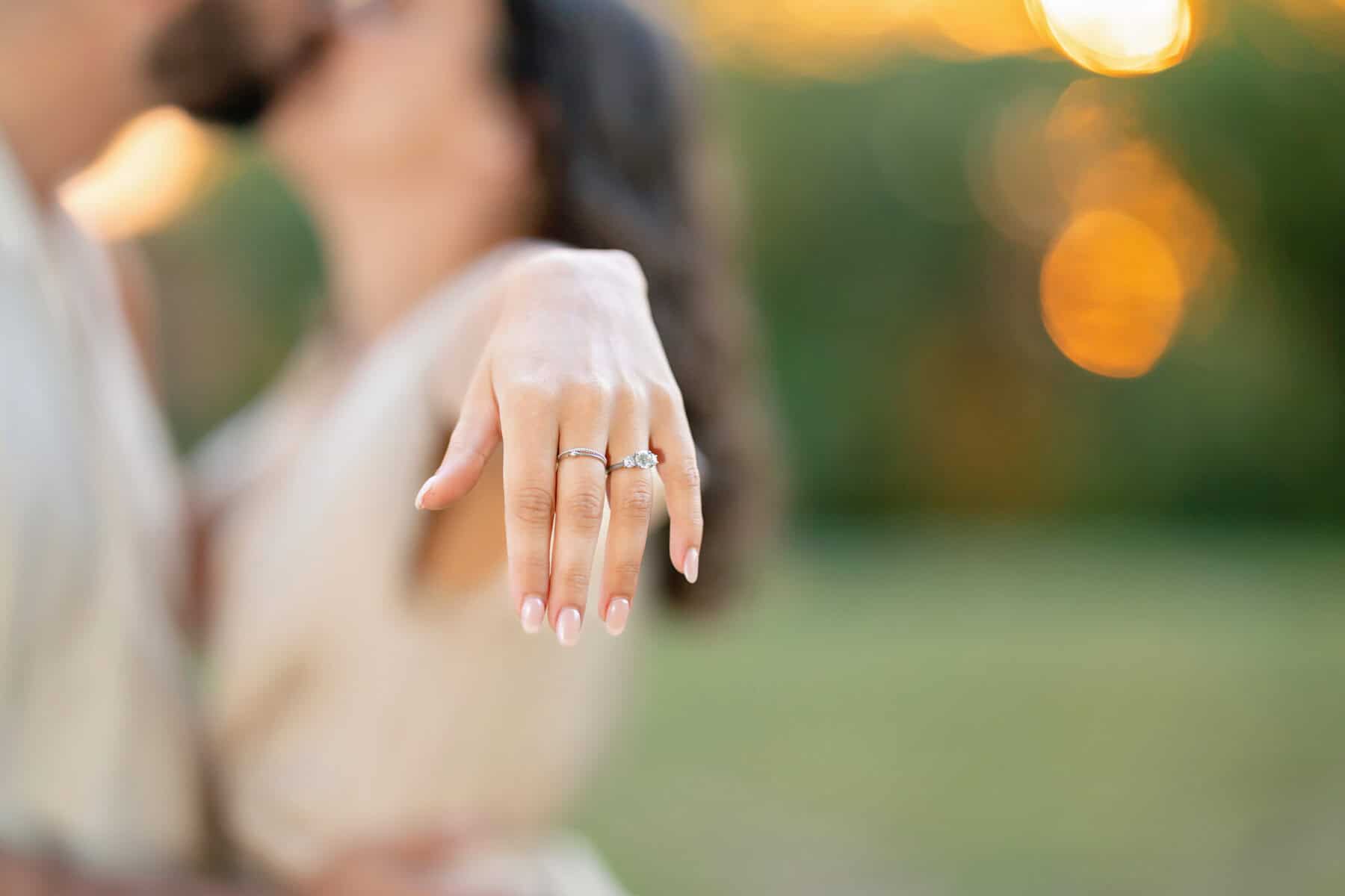 Webb Park in Weymouth, MA Engagement Session 17 A woman extends her hand to display an engagement ring during an Engagement Session at Webb Park, with her fiancé embracing her in the blurred sunset background.