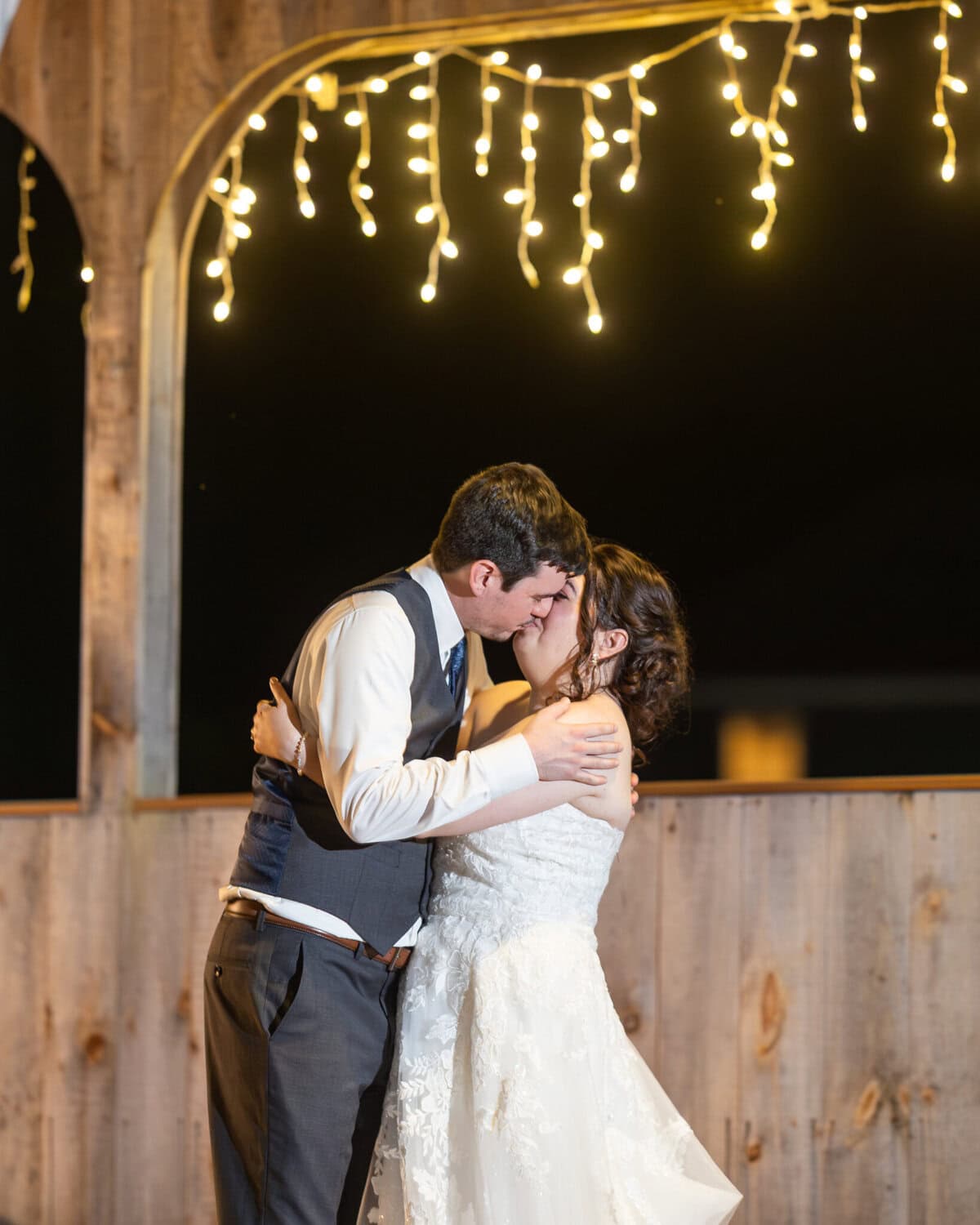 Stunning Wedding at Indian Ranch and Samuel Slater's Restaurant 109 A bride and groom share a kiss under string lights at night, celebrating their wedding by the wooden wall at Indian Ranch.