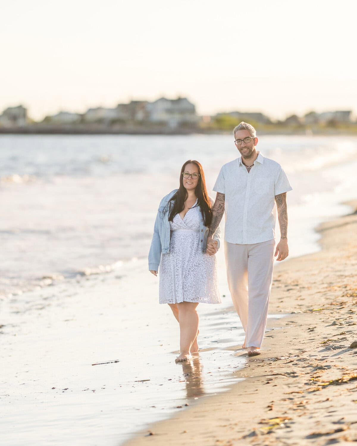 East Matunuck Beach Engagement Session 5 A couple enjoys an engagement session, walking barefoot along East Matunuck Beach at sunset, holding hands and smiling, with waves and beach houses softly blurred behind them.