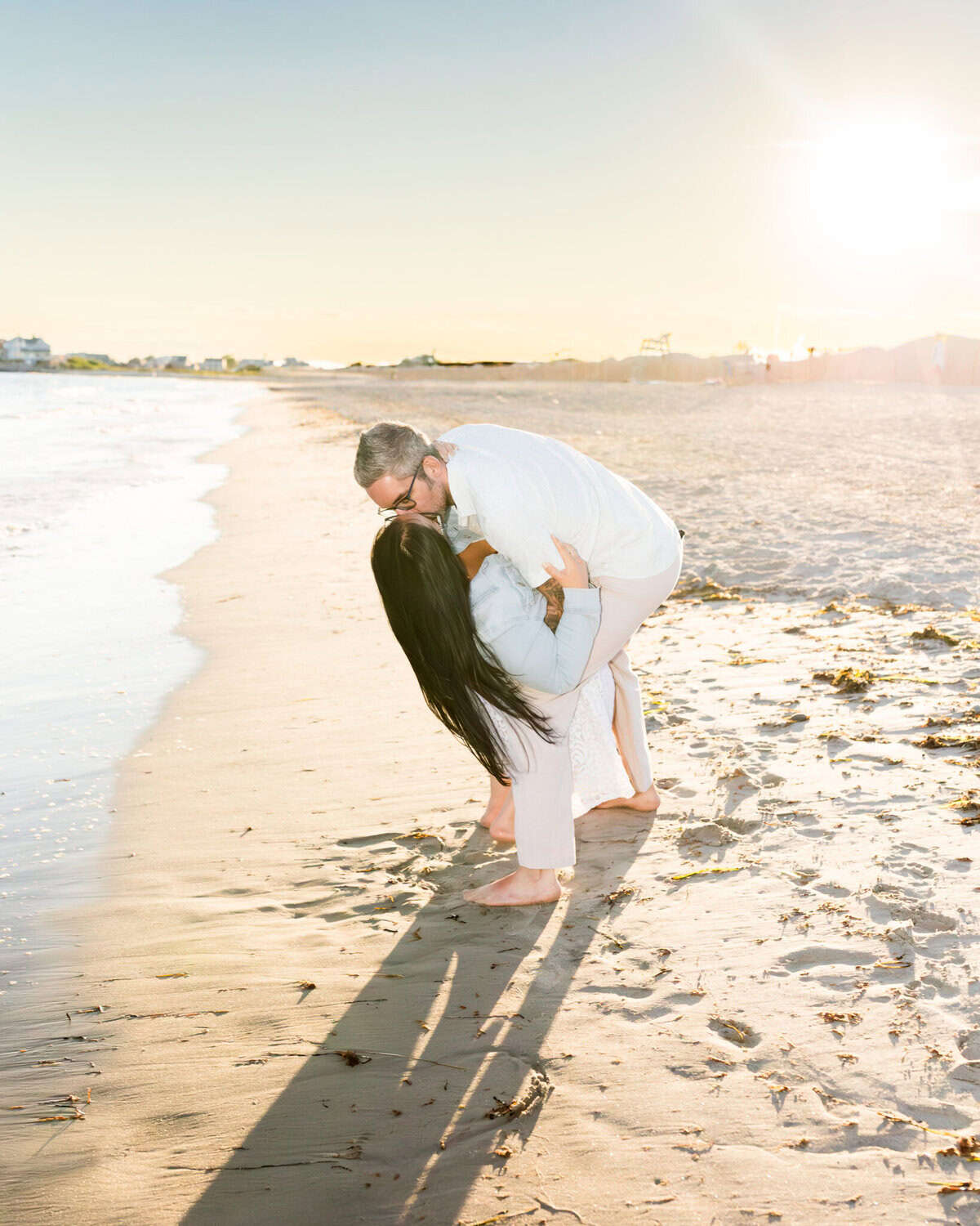 East Matunuck Beach Engagement Session 9 A couple in white shares a romantic dip and kiss during their Beach Engagement at East Matunuck Beach, casting long sunset shadows on the sand with the sun shining brightly behind them.