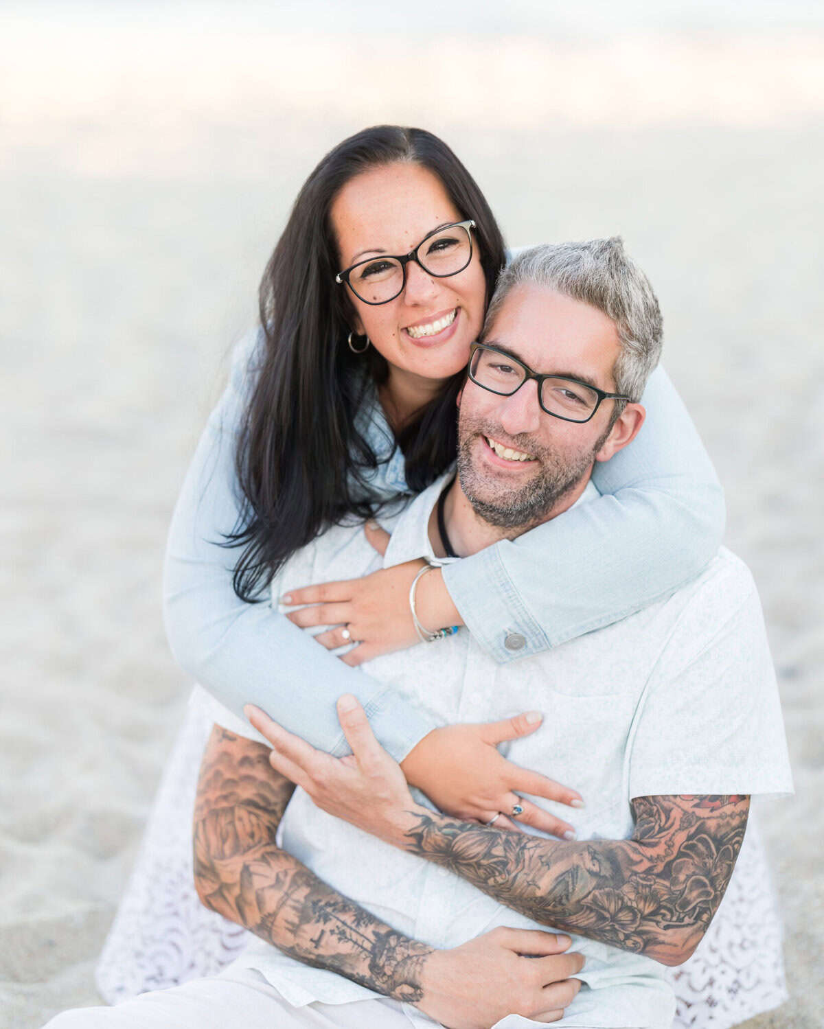 East Matunuck Beach Engagement Session 13 A smiling woman with long dark hair hugs a man with tattooed arms and glasses on East Matunuck Beach during their engagement session, both in light-colored clothing.
