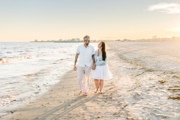 A man and woman walk barefoot, holding hands on East Matunuck Beach at sunset for an engagement session; gentle waves and golden sunlight complete the tranquil scene.
