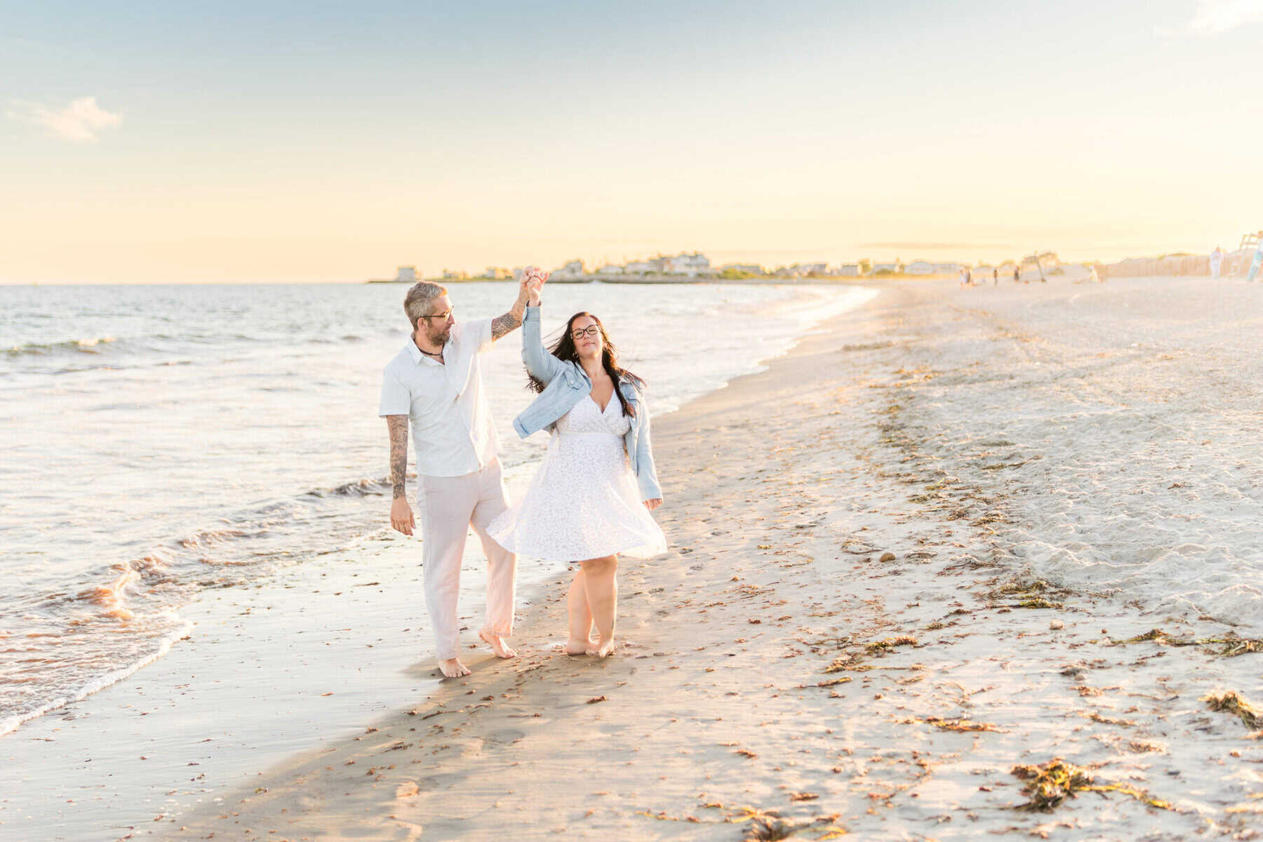 East Matunuck Beach Engagement Session 17 A smiling couple dances barefoot on East Matunuck Beach at sunset. The woman twirls in a white dress as the man holds her hand—perfect for a joyful beach engagement session.
