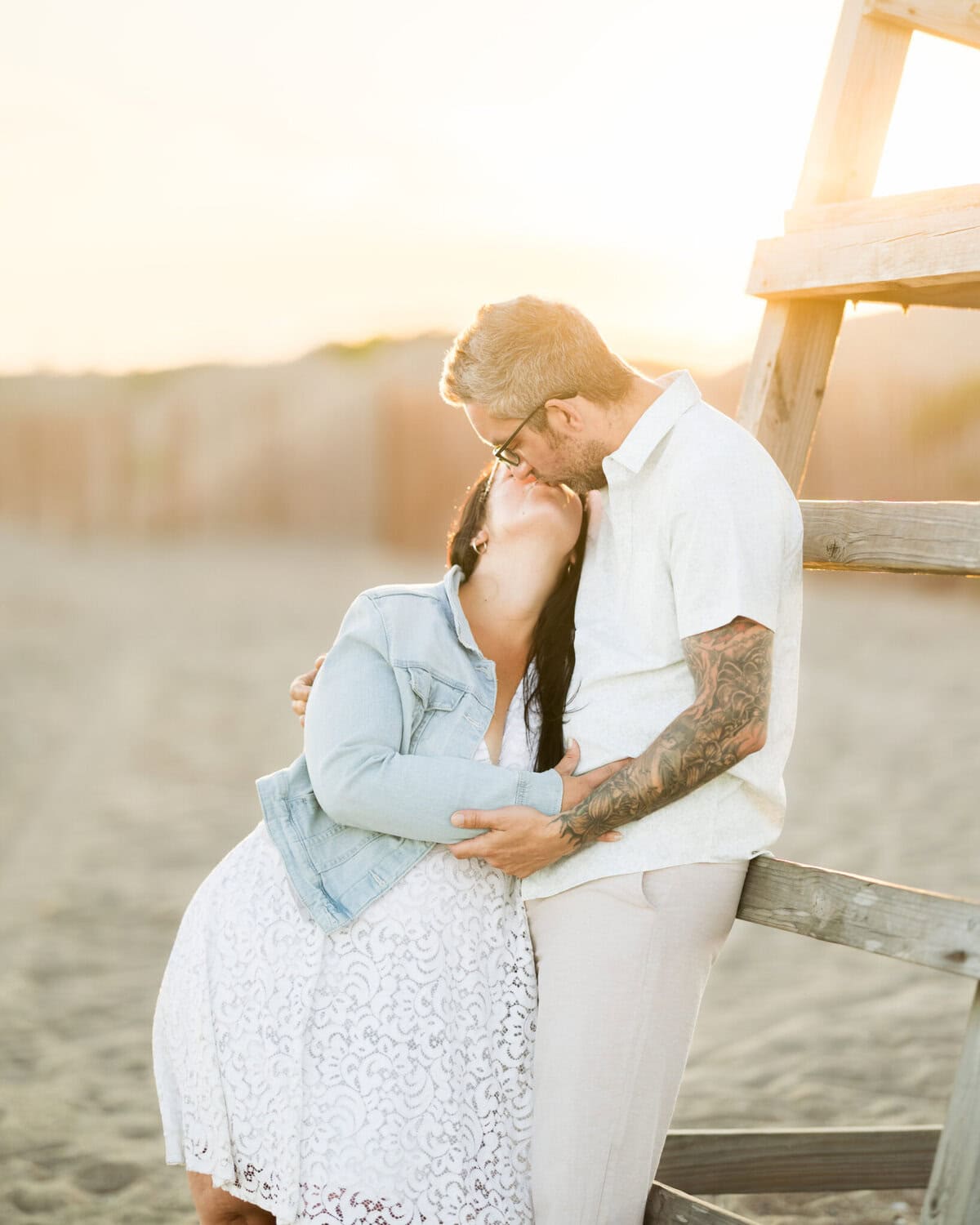 East Matunuck Beach Engagement Session 18 A couple shares a gentle kiss at East Matunuck Beach at sunset. The woman in a white dress and denim jacket leans toward her partner, making this engagement session feel warm and affectionate.