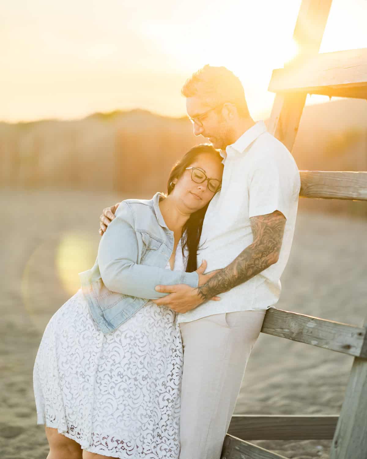 East Matunuck Beach Engagement Session 19 A couple embraces lovingly at East Matunuck Beach near a lifeguard stand at sunset, sharing a warm moment during their engagement session as sunlight glows softly behind them.