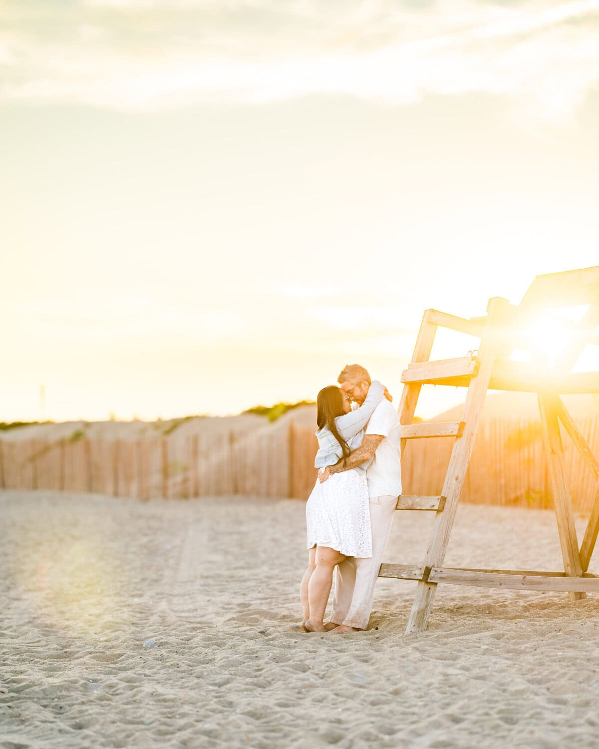 East Matunuck Beach Engagement Session 20 A couple embraces and kisses on sandy East Matunuck Beach near a lifeguard stand, sunlight glowing behind them—perfect for a romantic beach engagement session.