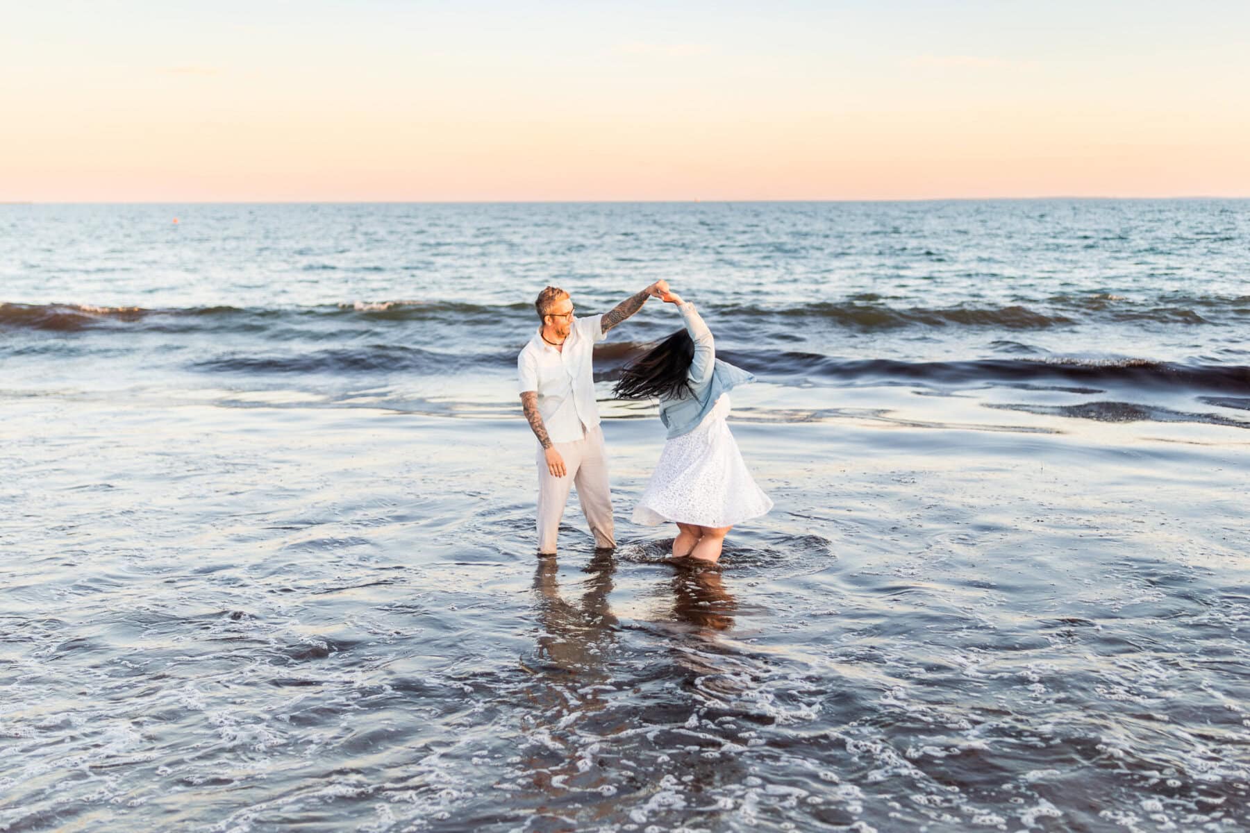 East Matunuck Beach Engagement Session 21 A couple dances in shallow ocean water at sunset during their Beach Engagement; the man twirls his partner as waves roll in and a pastel sky glows behind them at East Matunuck Beach.