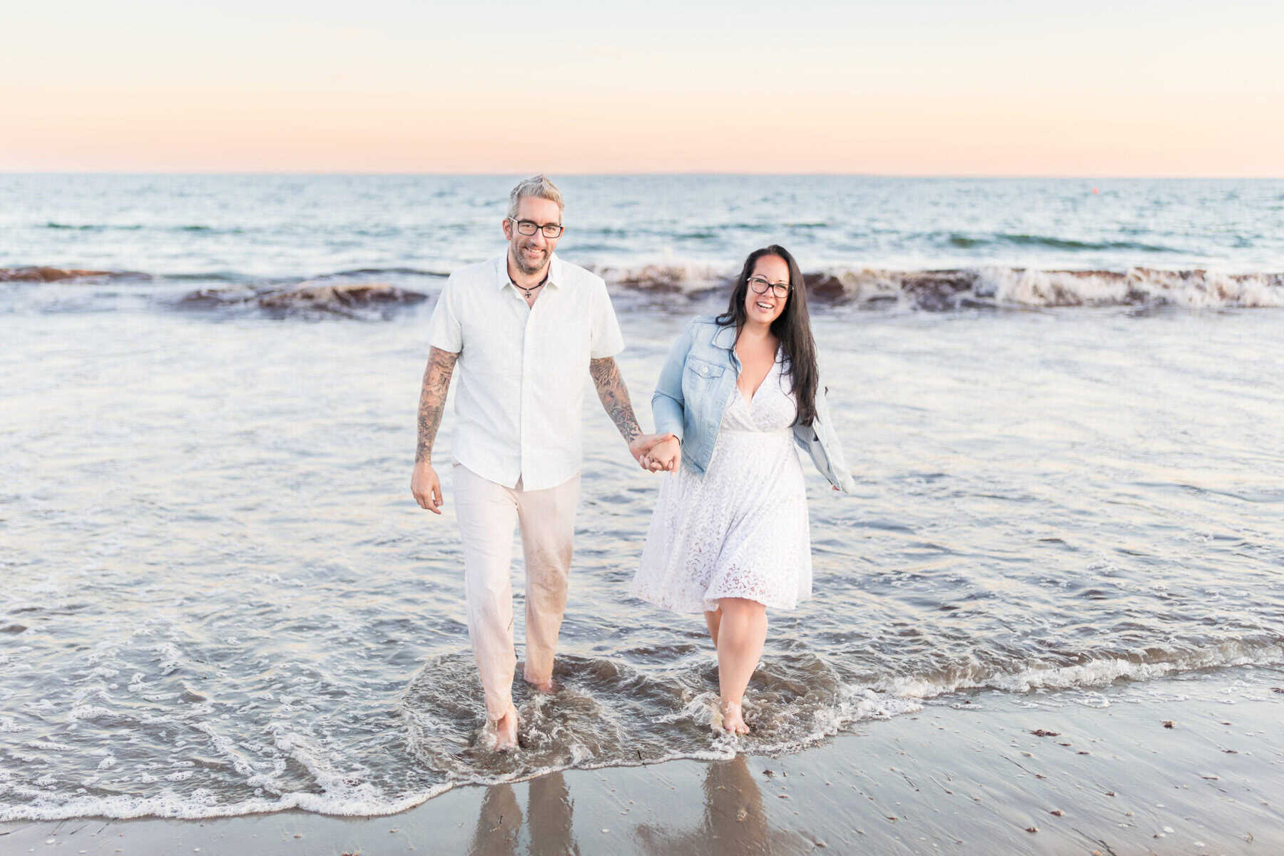 East Matunuck Beach Engagement Session 24 A smiling couple walks barefoot on East Matunuck Beach for their engagement session, holding hands as gentle waves and a pastel sunset sky create the perfect beach engagement backdrop.