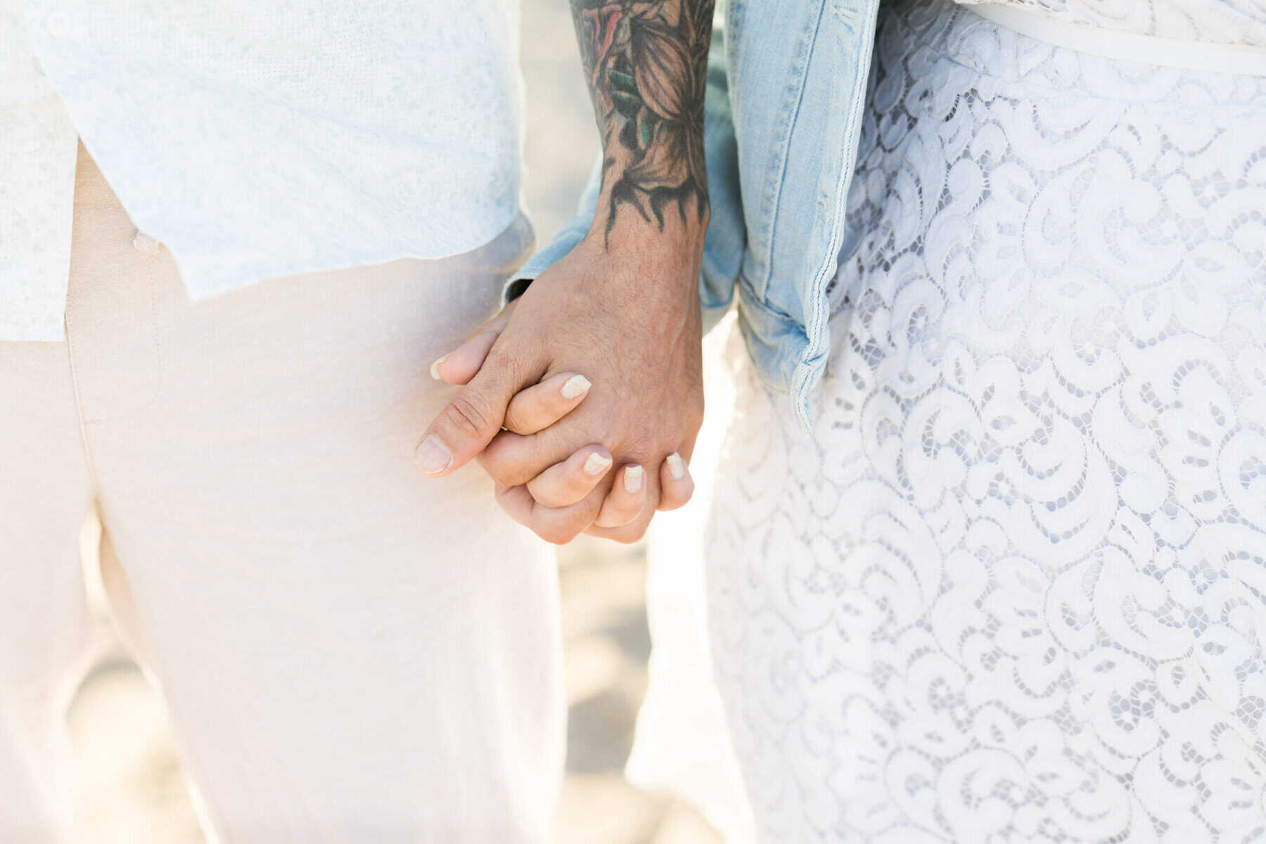East Matunuck Beach Engagement Session 25 During this East Matunuck Beach engagement session, two people with light skin and manicured nails hold hands—one in a white lace dress, the other with a tattooed arm and light pants.