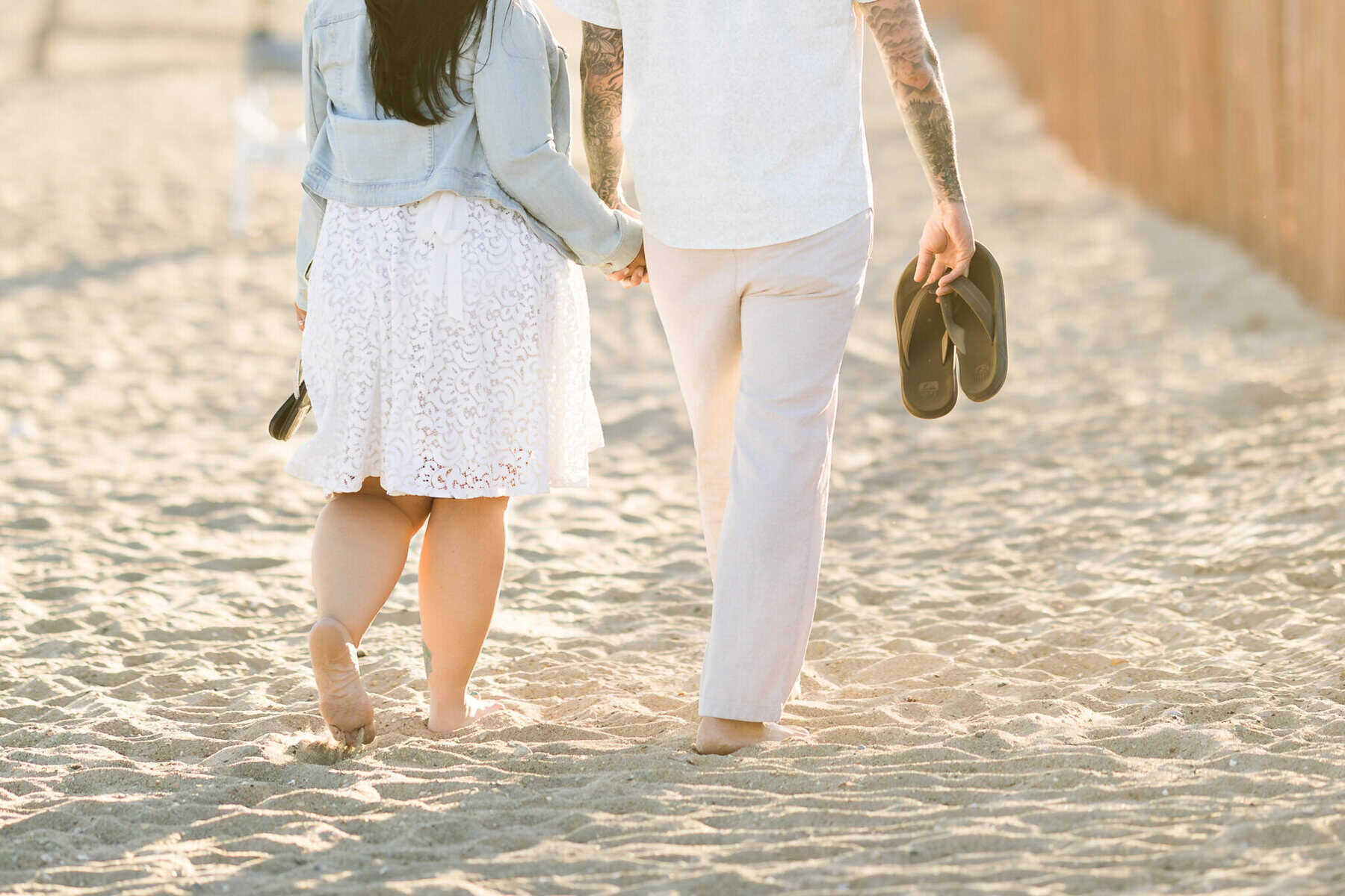 East Matunuck Beach Engagement Session 27 A couple walks barefoot on East Matunuck Beach, holding hands. The woman wears a white dress and jean jacket; he holds sandals. Sunlight creates a warm, soft engagement session glow.