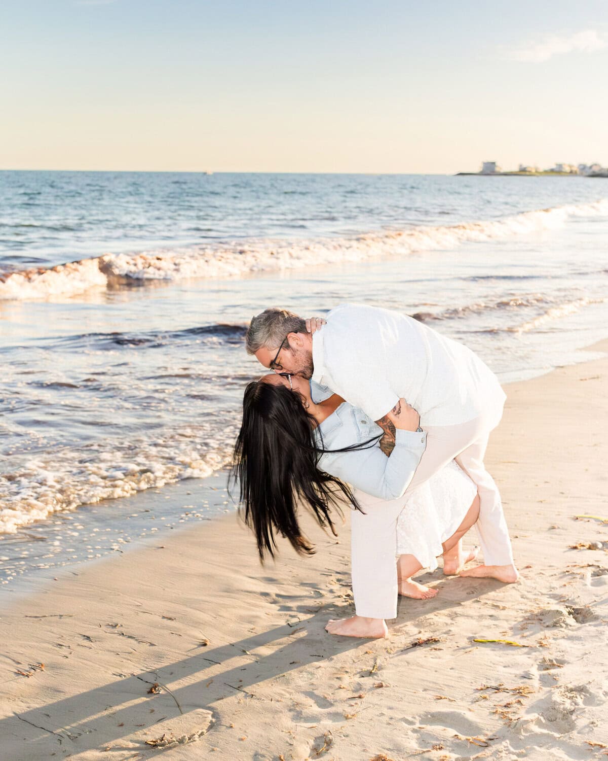 East Matunuck Beach Engagement Session 2 A man in white clothes dips and kisses a woman with long dark hair on East Matunuck Beach at sunset—an affectionate moment captured in this joyful engagement session.