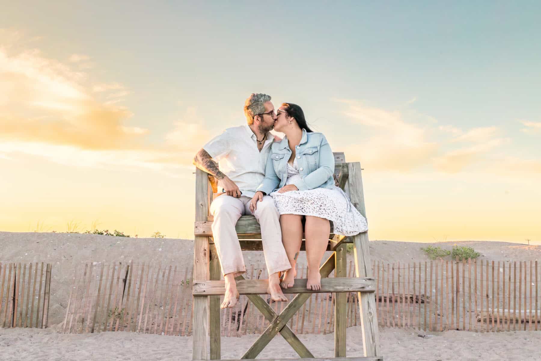 East Matunuck Beach Engagement Session 4 A couple sits barefoot on a lifeguard stand at East Matunuck Beach, kissing at sunset. Their light clothes and relaxed smiles capture the joy of this engagement session.