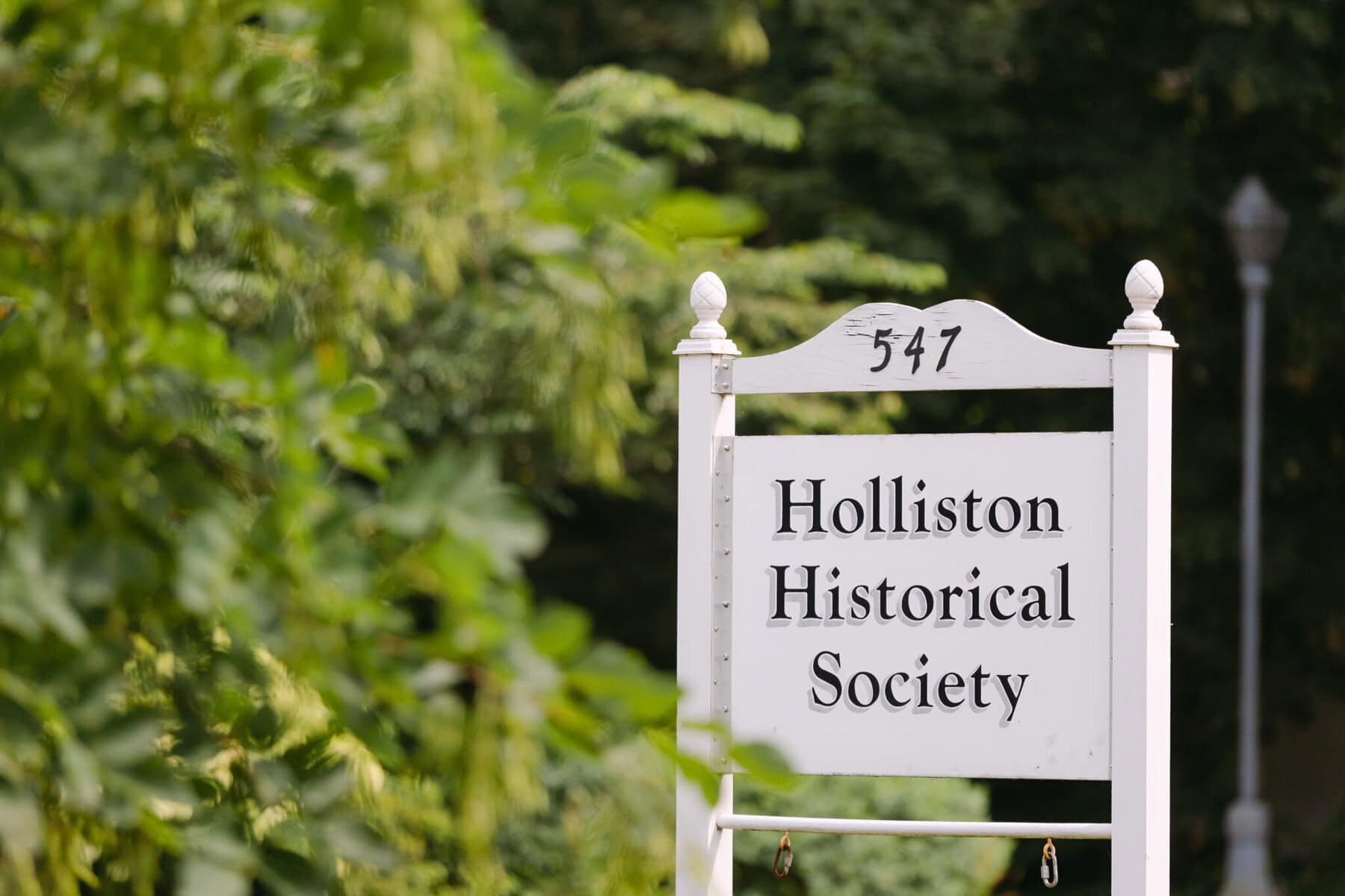 Wedding At the Holliston Historical Society 2 White wooden sign reads "547 Holliston Historical Society," a charming wedding venue, surrounded by lush green foliage and a blurred background.