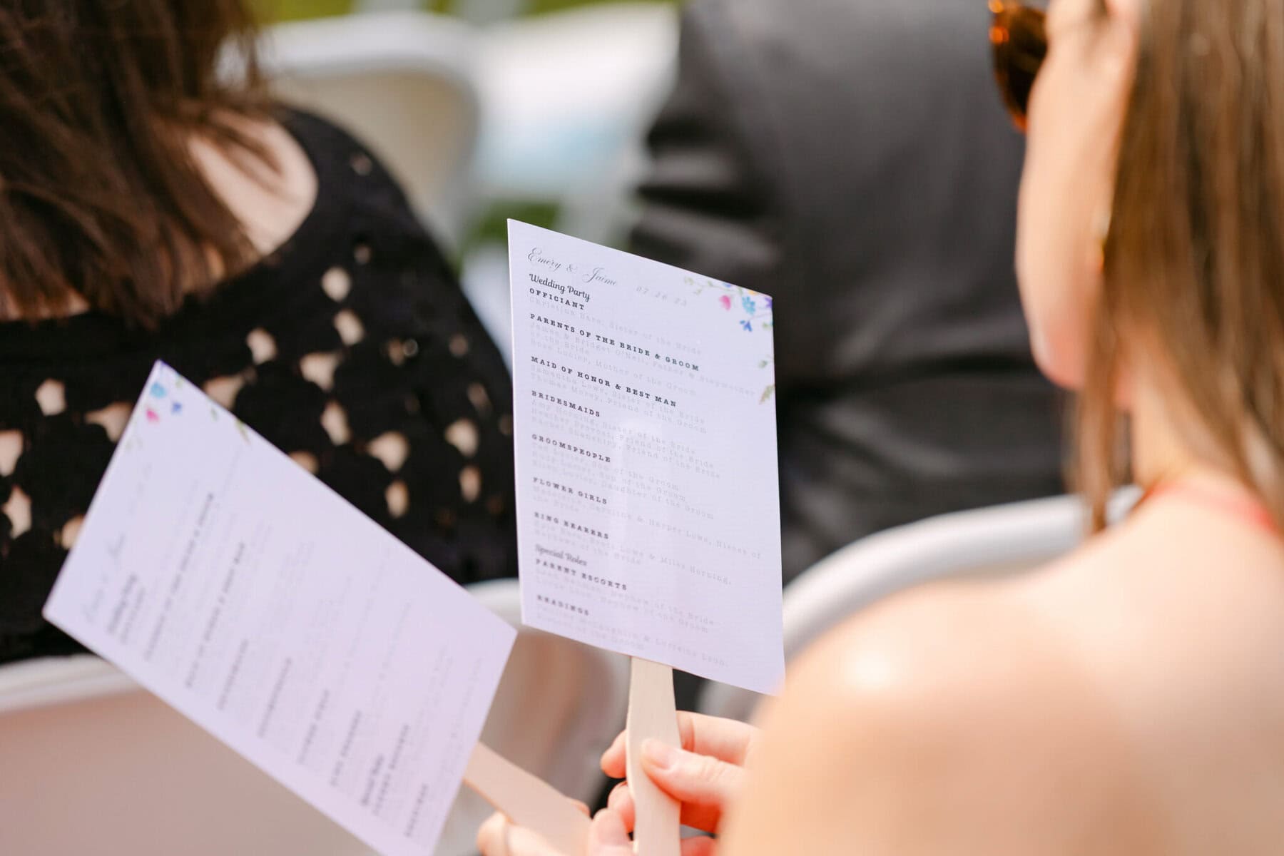 Wedding At the Holliston Historical Society 5 A person holds a printed wedding program on a stick, seated among others at an outdoor event at Holliston Historical Society.