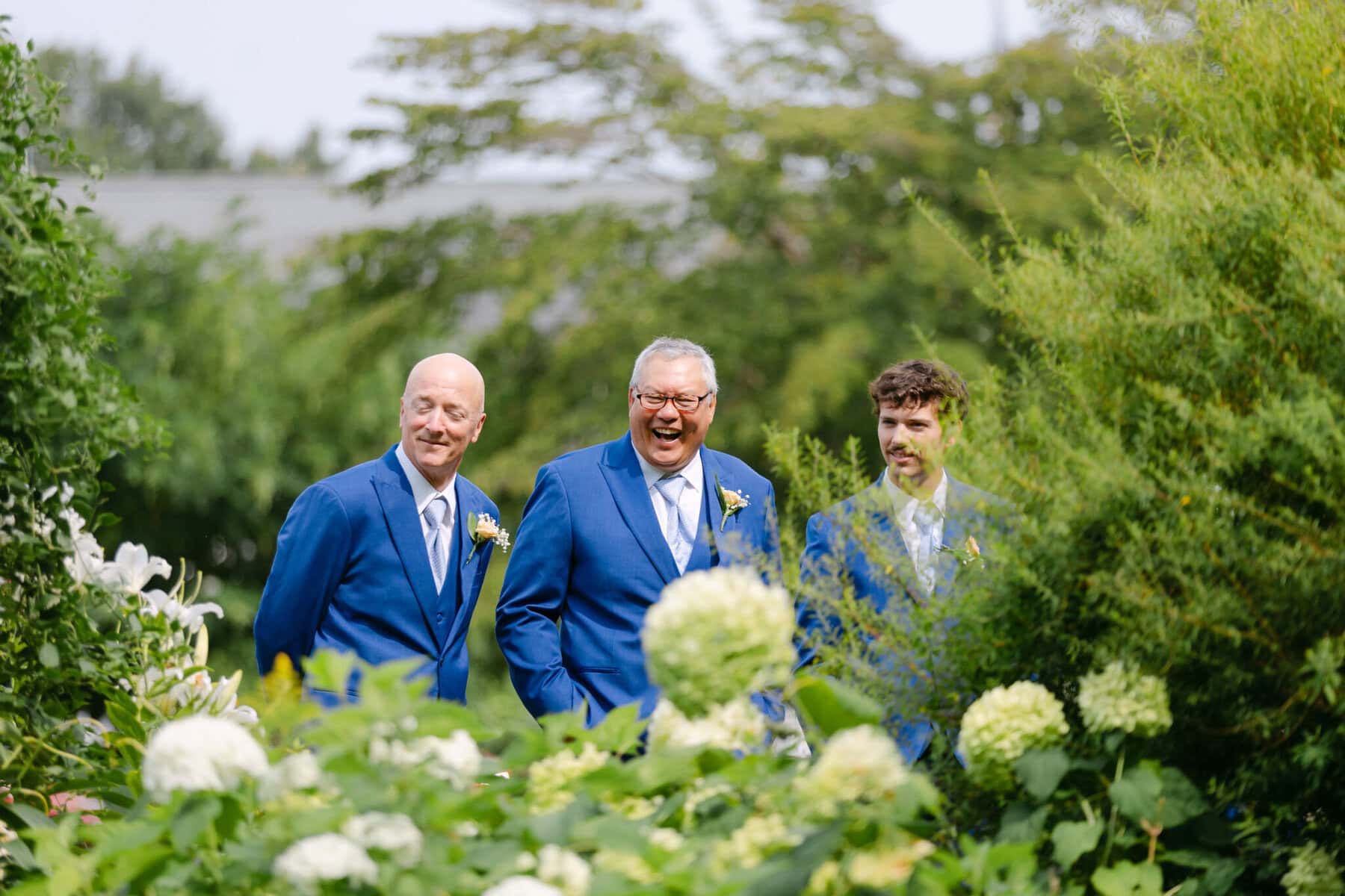 Wedding At the Holliston Historical Society 7 Three men in blue suits with boutonnieres laugh together at a wedding in the garden of the Holliston Historical Society.