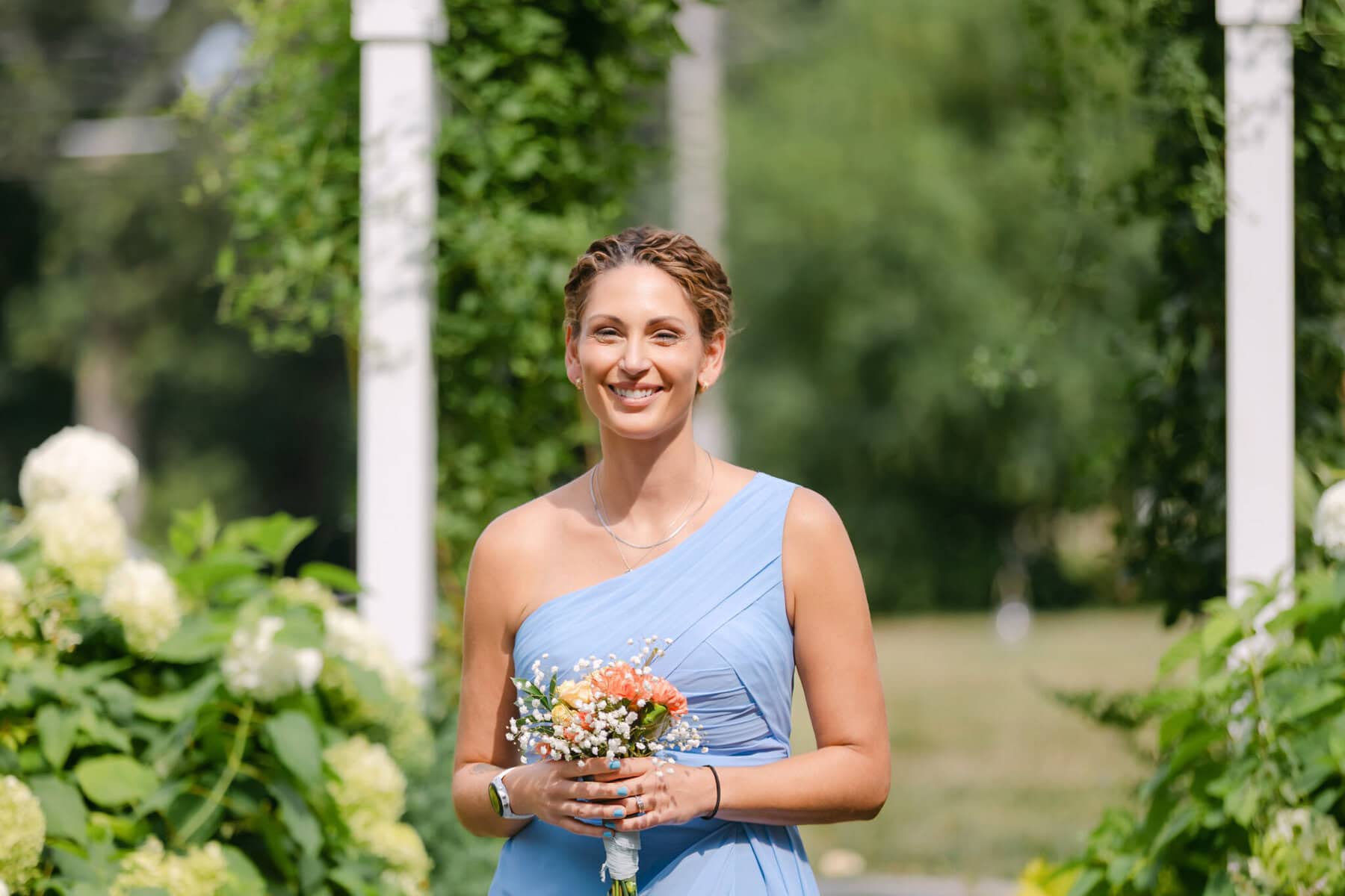 Wedding At the Holliston Historical Society 9 Woman in a light blue, one-shoulder dress holds a bouquet at a wedding venue among greenery and blooms at Holliston Historical Society.