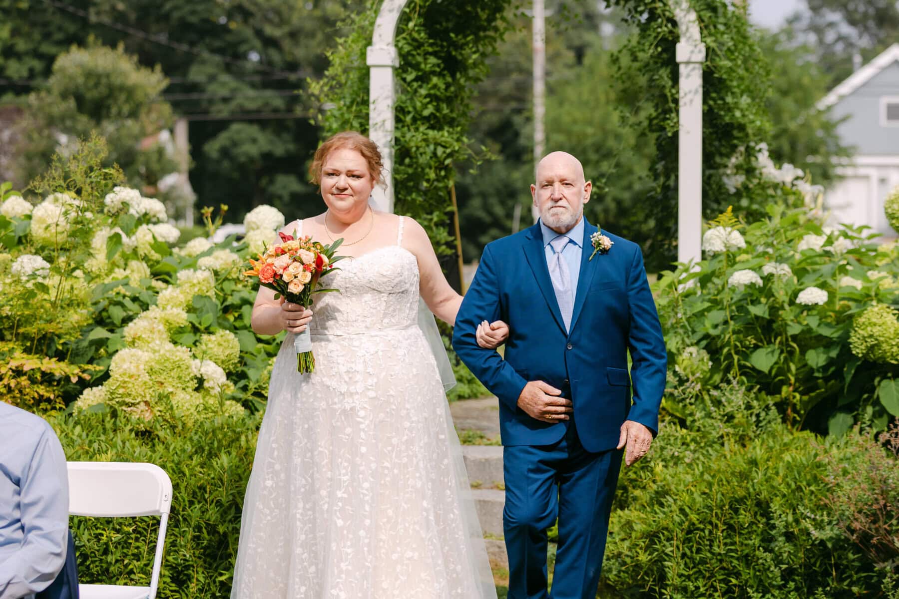 Wedding At the Holliston Historical Society 13 A bride holding a bouquet walks outside with an older man at the Holliston Historical Society wedding venue, surrounded by lush greenery.