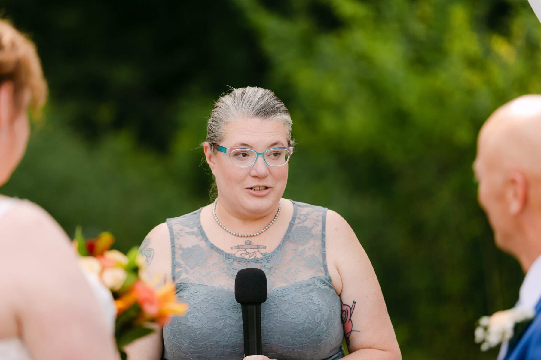 Wedding At the Holliston Historical Society 16 A person in a gray lace dress holds a microphone, speaking at a Wedding outdoors at the Holliston Historical Society.