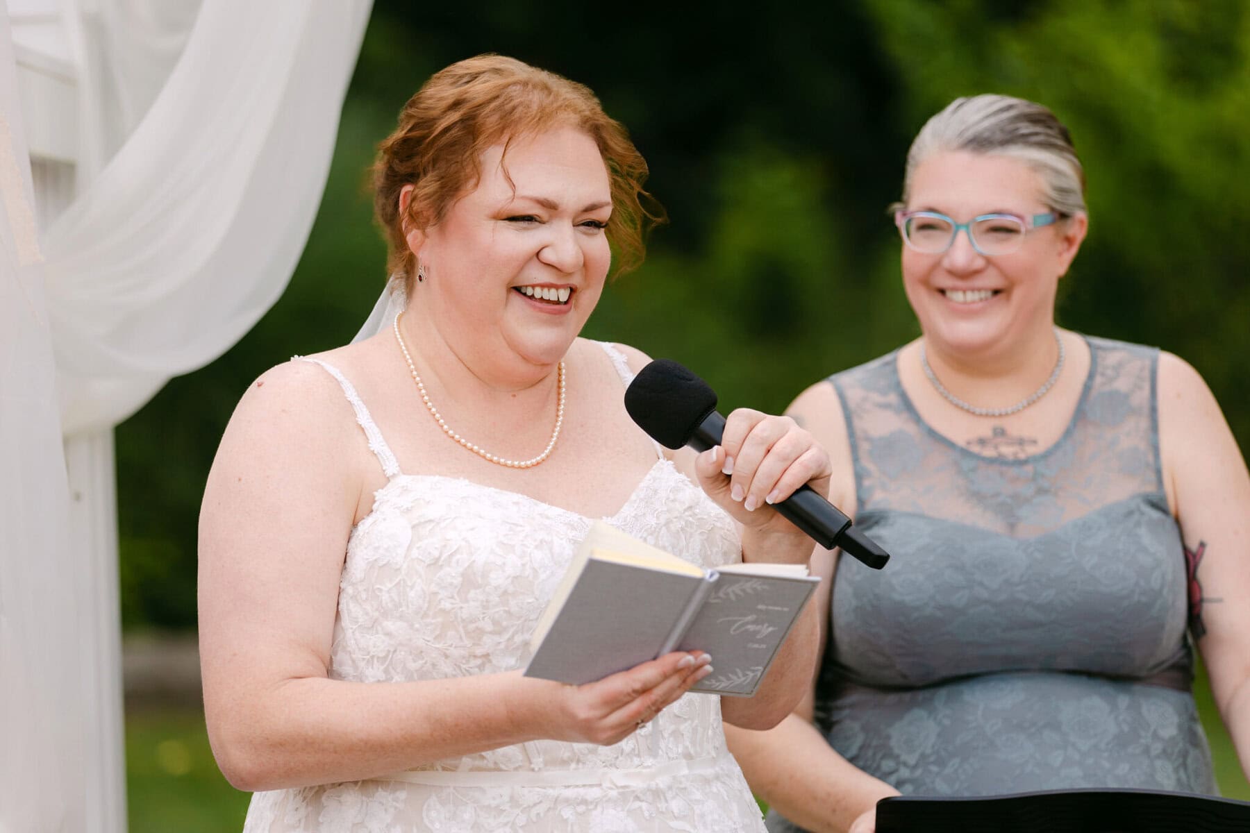 Wedding At the Holliston Historical Society 17 A woman in a white dress holds a microphone and notebook, speaking at a wedding at the Holliston Historical Society beside a smiling guest.