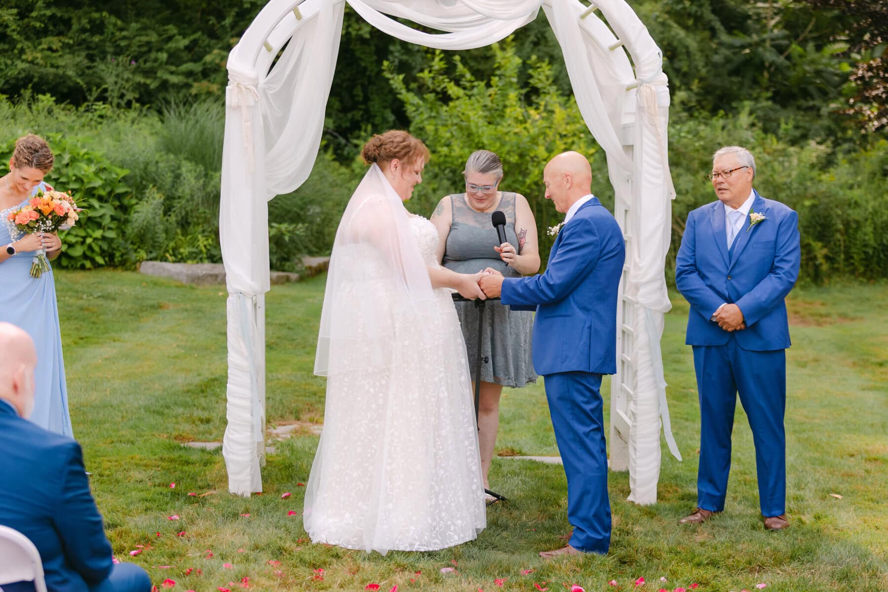 Wedding At the Holliston Historical Society 22 A bride and groom hold hands under a white arch at the Holliston Historical Society wedding venue, joined by an officiant and two attendants.