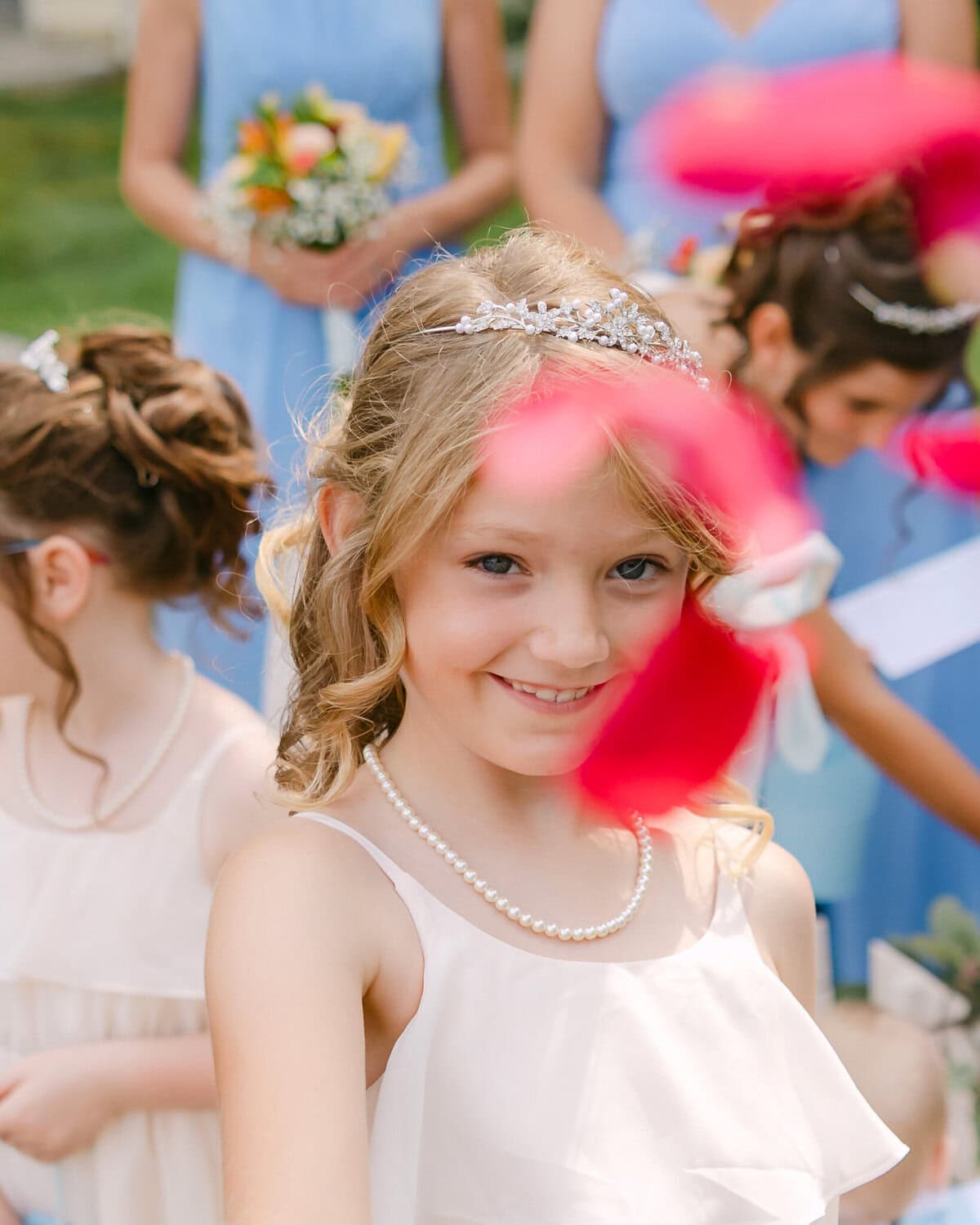 Wedding At the Holliston Historical Society 11 Young girl with curled hair, tiara, and pearls smiles at a wedding venue; red petals in foreground, others stand behind her.