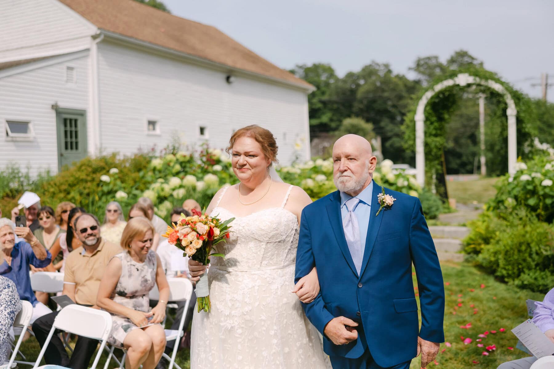 Wedding At the Holliston Historical Society 14 A bride holding a bouquet walks down the aisle with an older man in a blue suit at the Holliston Historical Society wedding venue.