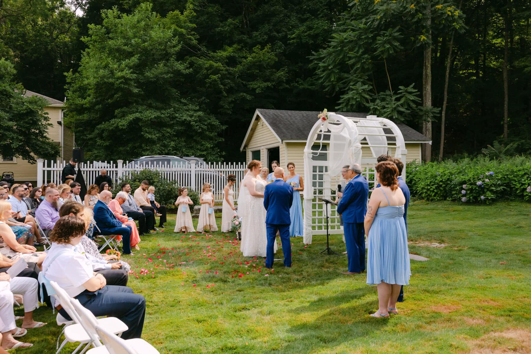 Wedding At the Holliston Historical Society 21 A bride and groom stand under a white arbor at the Holliston Historical Society wedding venue, surrounded by guests and the wedding party.