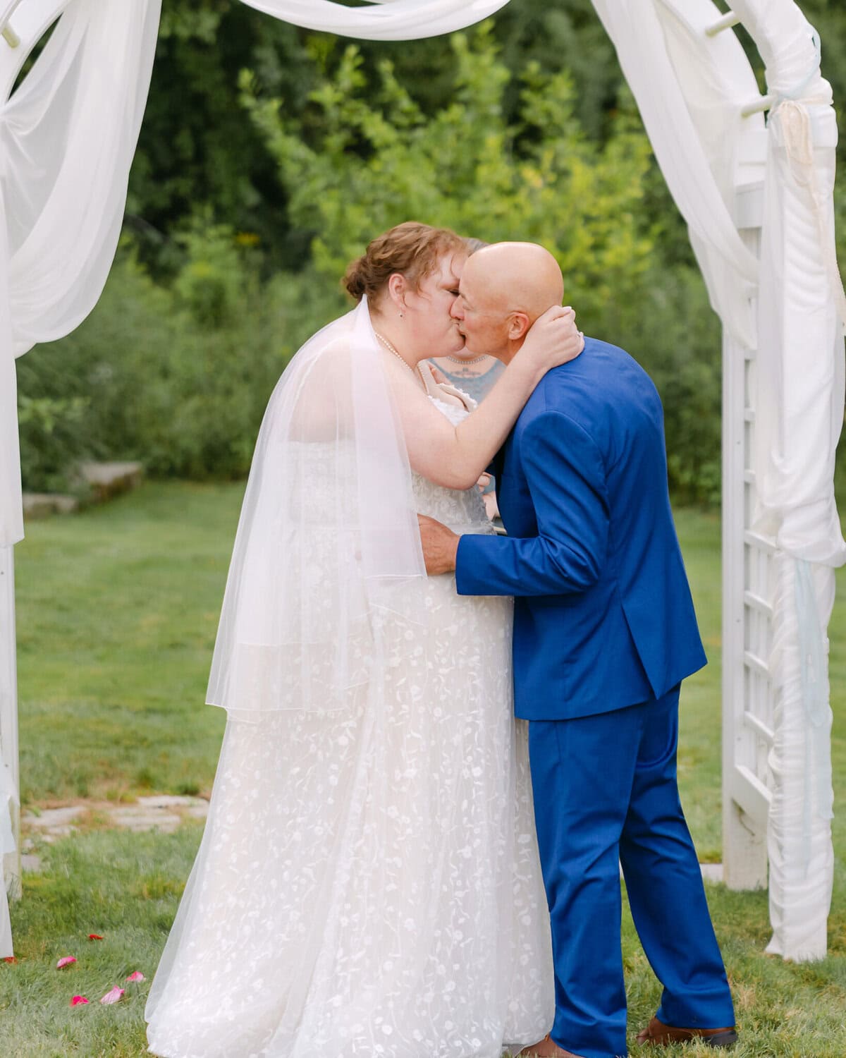 Wedding At the Holliston Historical Society 26 A bride and groom kiss under a white arch at the Holliston Historical Society wedding venue, surrounded by lush greenery.