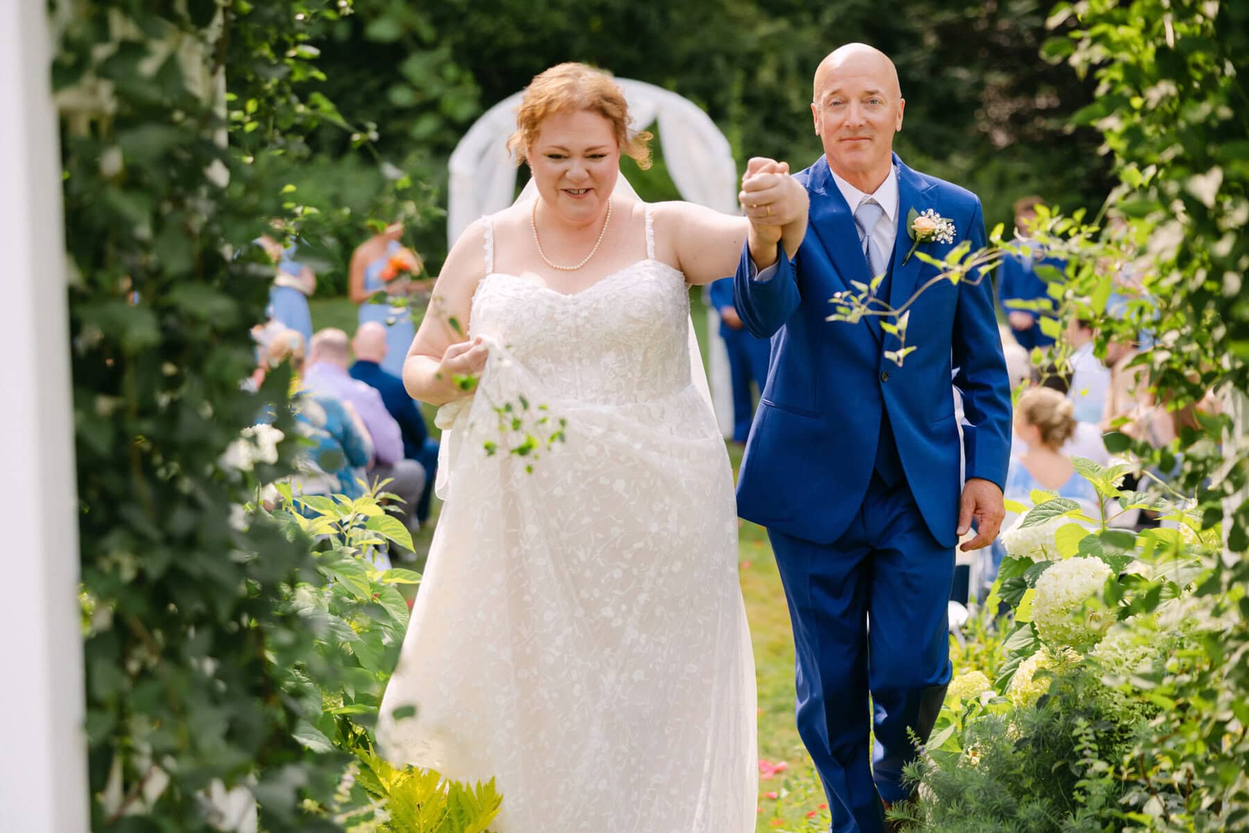 Wedding At the Holliston Historical Society 28 A bride in a white dress and groom in blue suit walk hand-in-hand at their wedding at Holliston Historical Society.