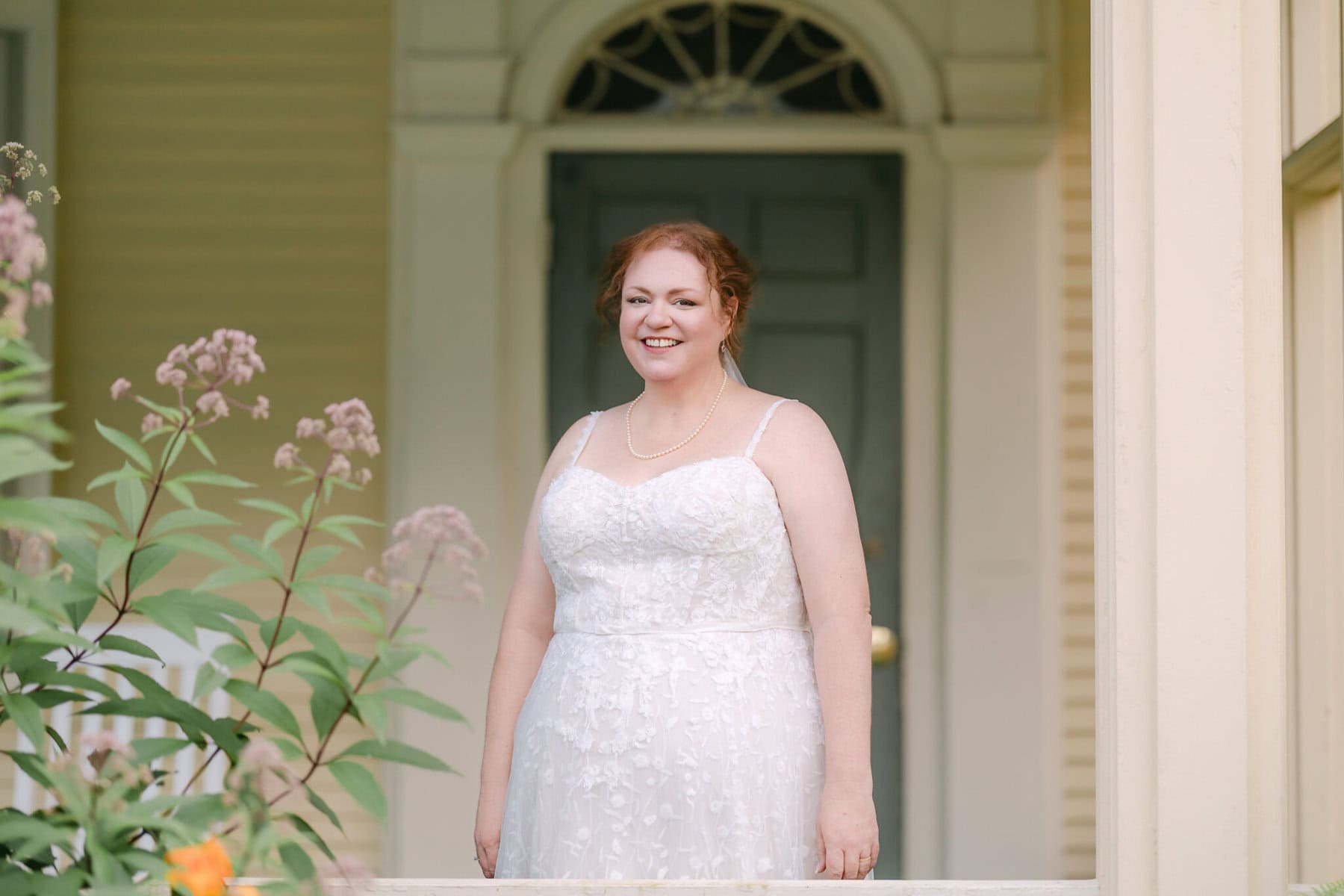 Wedding At the Holliston Historical Society 53 A woman in a white lace wedding dress stands on a porch at Holliston Historical Society, smiling amid greenery and flowers.