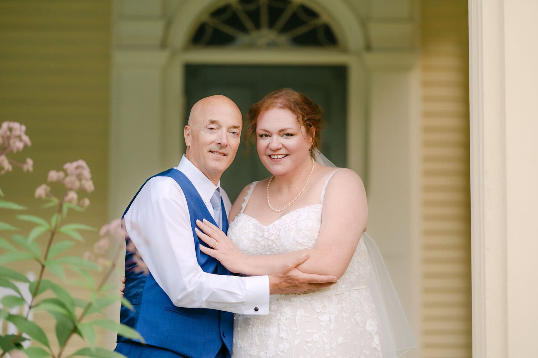 Wedding At the Holliston Historical Society 54 A man in a blue vest and a woman in a white wedding dress pose together outside the Holliston Historical Society house.