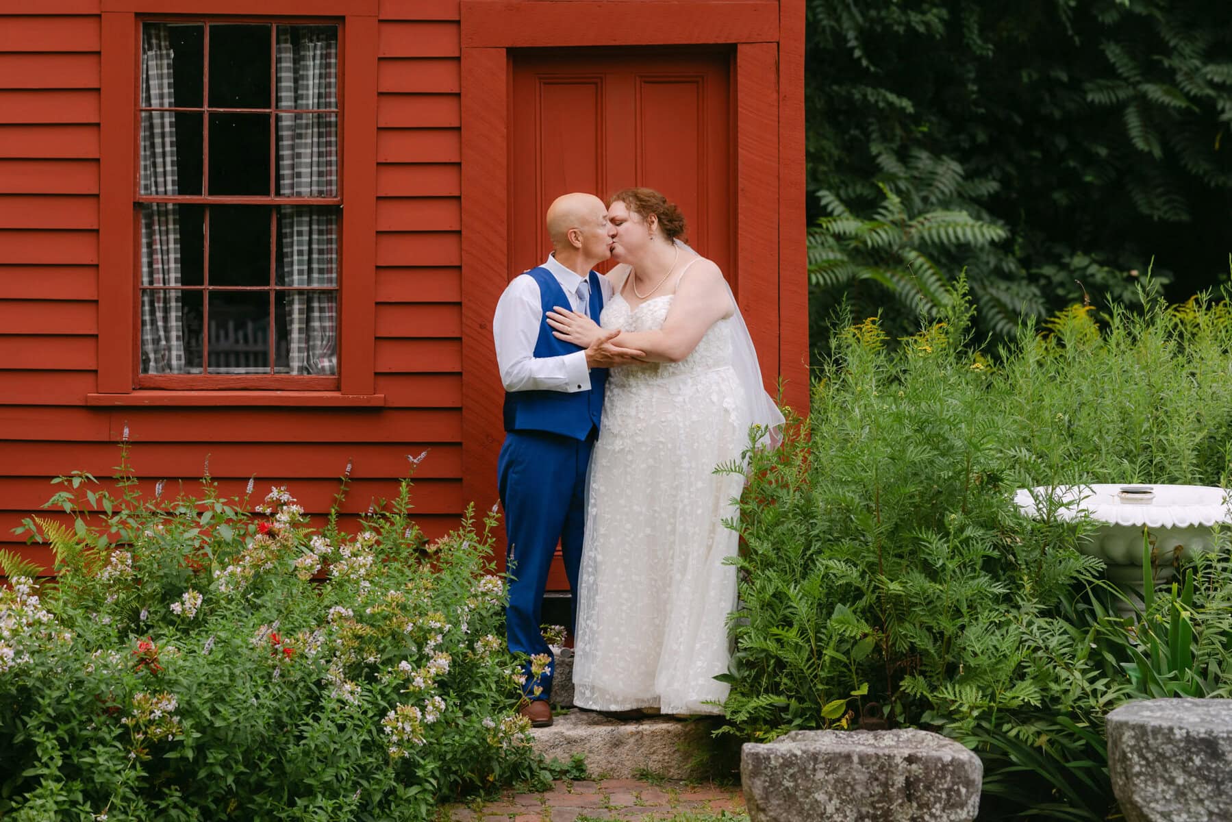 Wedding At the Holliston Historical Society 56 A couple in wedding attire embraces and smiles in front of a red wooden building at the Holliston Historical Society, surrounded by greenery and flowers.