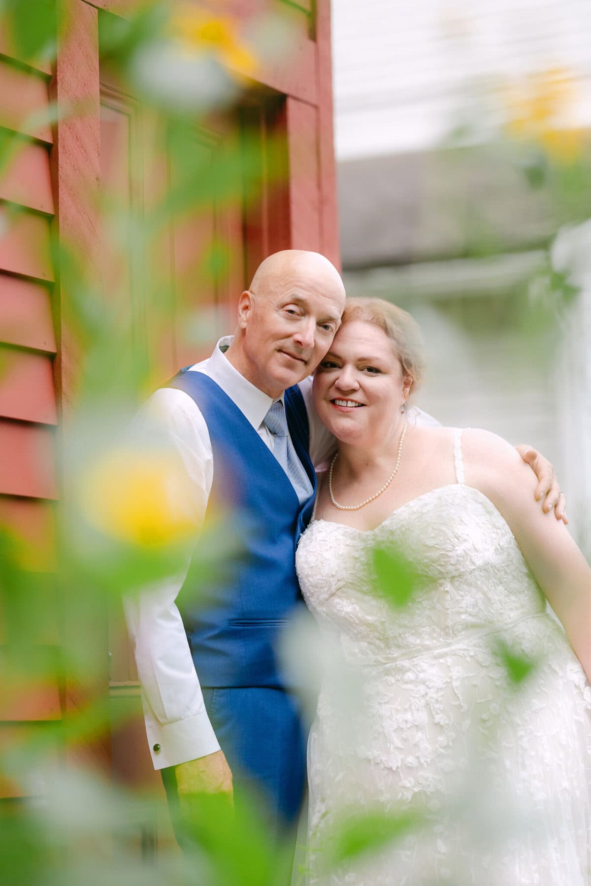 Wedding At the Holliston Historical Society 57 A couple in wedding attire stands outside the Holliston Historical Society, framed by blurred green plants and yellow flowers.