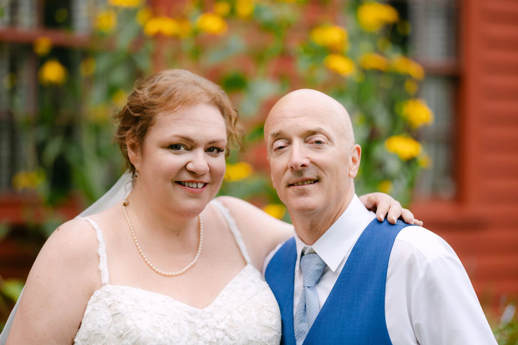 Wedding At the Holliston Historical Society 59 A bride and groom smile outside at the Holliston Historical Society, yellow flowers in bloom, capturing a joyful wedding moment.